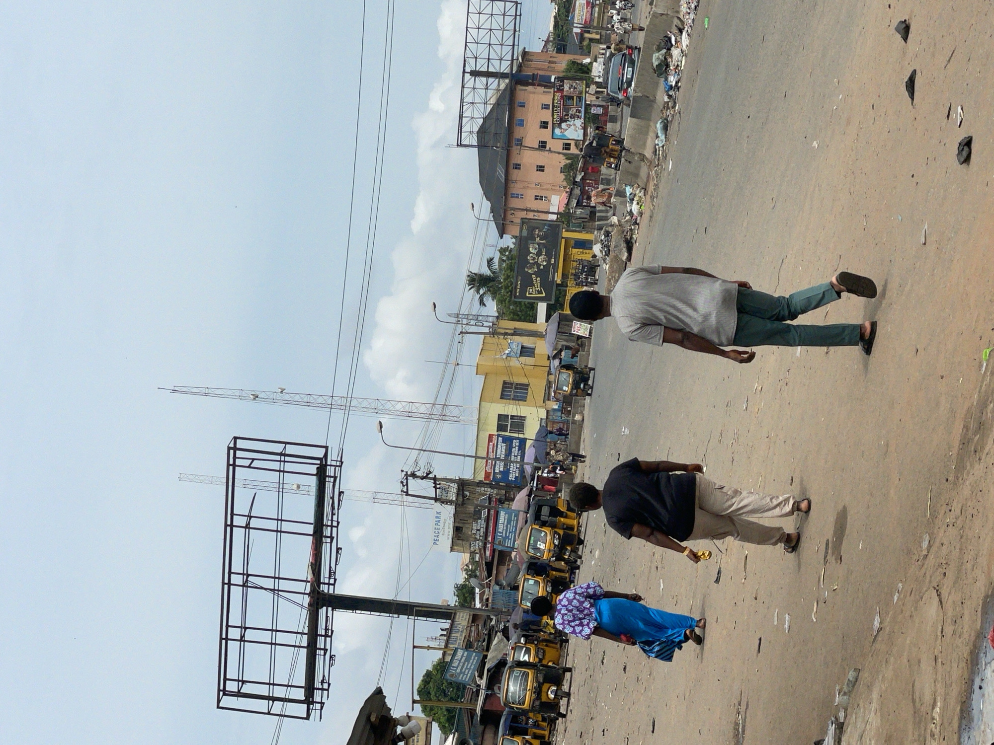 The image shows a street scene in Nkpor, Nigeria. Three people are walking on the side of a road. The road has visible litter and trash along the side. Several vehicles, particularly yellow minibuses (tricycles or "keke napep"), are present on the road. Above are billboards and antennas. There is a building in the background. The sky is overcast.