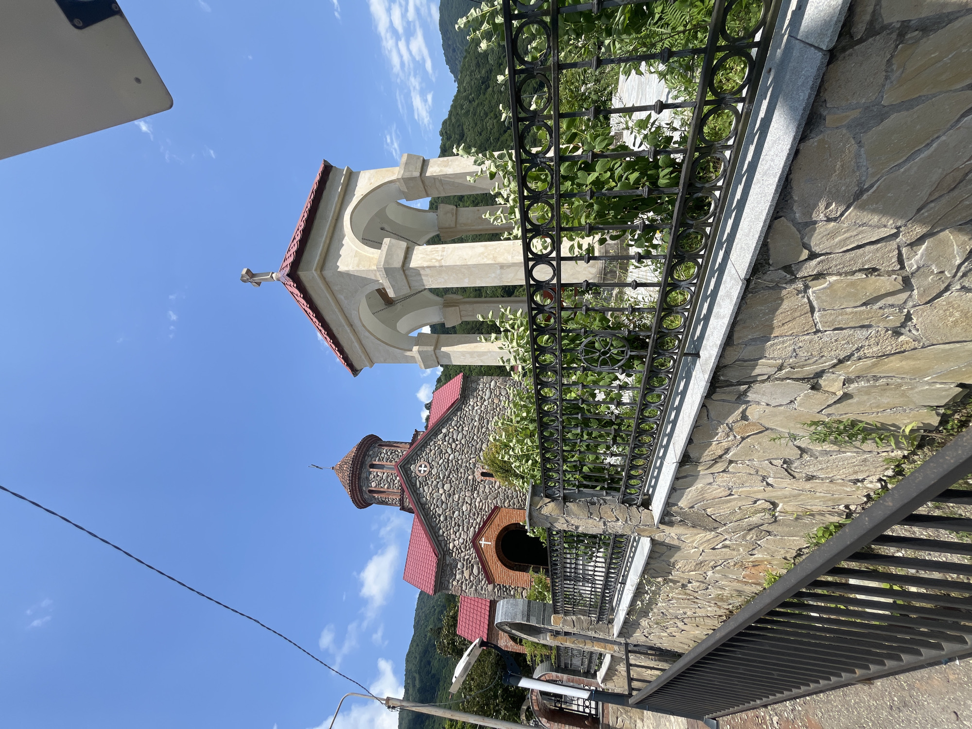 A medium shot captures a stone church with a red tiled roof in Adjaristskali, Georgia. To the right of the church is a light-colored, open-air bell tower with a red tiled roof and a cross at its apex. Both the church and bell tower are partially obscured by a black metal fence and lush green foliage. The church is constructed of rough-hewn stones, with a prominent arched entranceway. The bell tower features four arched openings supported by stone pillars. Behind the structures, a verdant, tree-covered mountainside rises under a clear blue sky dotted with a few white clouds. A stone wall runs along the foreground, topped by the black metal fence. A narrow asphalt pathway or road is visible to the left, bordered by a black metal railing. A utility pole with a light fixture stands to the left of the church.