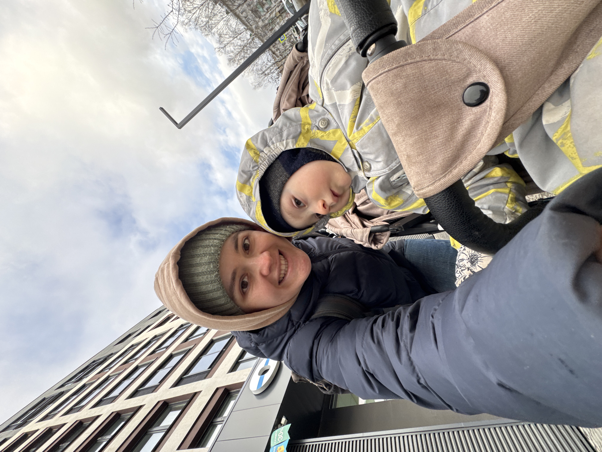 The image captures a tilted, low-angle selfie of a smiling woman and a young child, likely a mother and her baby or toddler, outdoors in Izhevsk, Russia, on an overcast day. Both subjects are dressed warmly in winter attire. The woman, on the left, wears a dark blue puffer jacket and a green knitted hat beneath a beige hood, smiling broadly at the camera. The child, seated in a stroller on the right, wears a grey and yellow patterned winter jacket with a hood over a dark beanie, looking directly at the lens with a neutral, slightly curious expression.
The background features a modern building with distinctive architectural elements, including numerous angular windows and alternating light and dark facade panels. A blue circular logo is visible on the building's exterior, beneath which a yellow sign displays Cyrillic text, including "СКИДКИ" (Discounts) and "30%". The sky is cloudy, and bare tree branches are visible towards the top. A metal L-shaped structure protrudes into the frame from the top right, and a grated surface appears at the bottom right, suggesting an urban setting. The overall impression is a cheerful moment shared between parent and child during an outing.