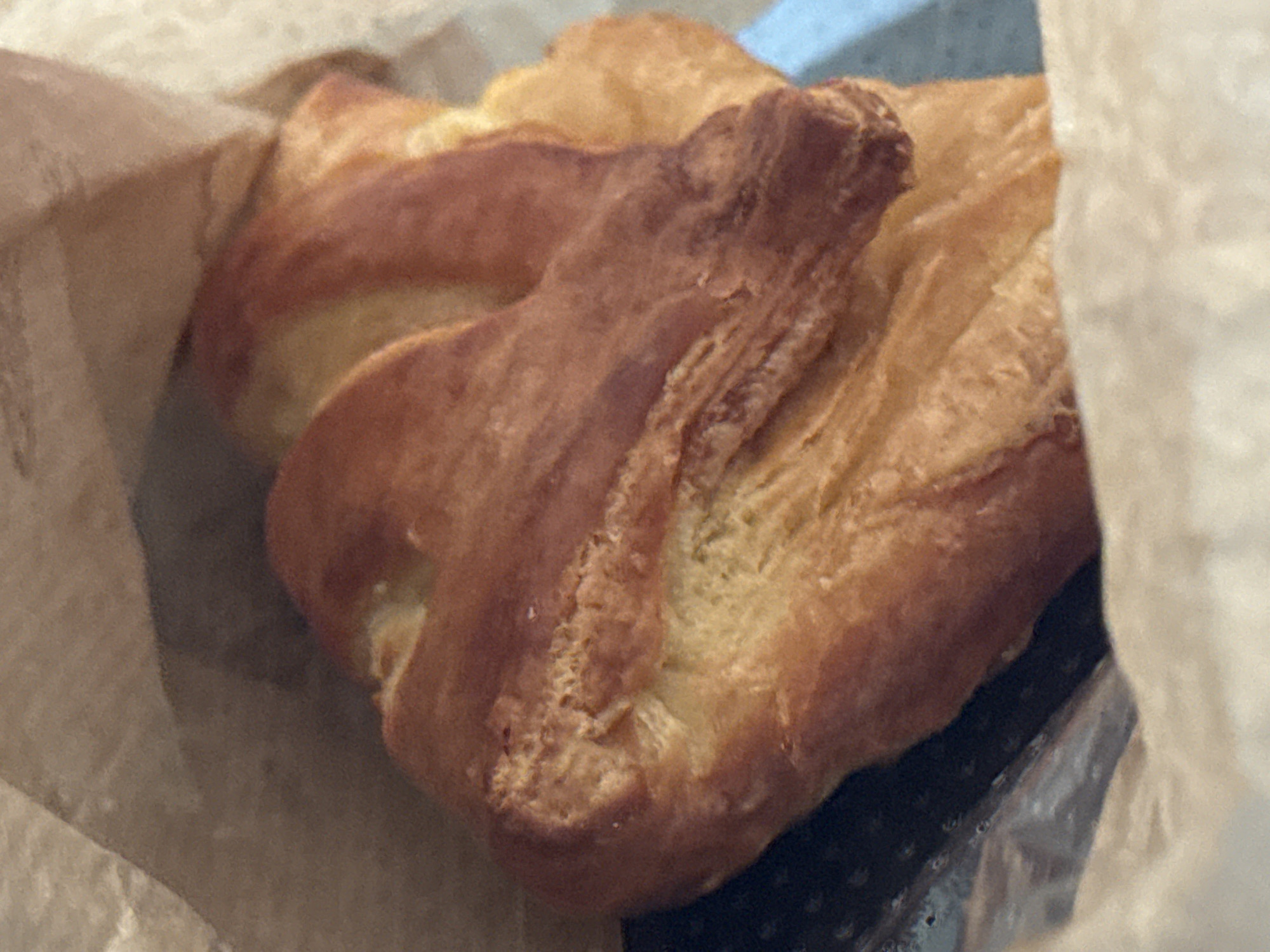 A close-up, eye-level shot captures a golden-brown croissant resting inside a brown paper bag. The croissant, which appears to be flaky and layered, is positioned in the center of the frame, taking up most of the space. Its surface exhibits a rich, baked texture with some darker brown hues indicating where it is more browned. The paper bag is visible surrounding the pastry, with its crinkled texture adding a sense of depth. The lighting is soft, casting gentle shadows and highlighting the croissant's golden color. The background is a blur of muted tones, suggesting an indoor setting, potentially a bakery or kitchen. There are no people visible in the image.