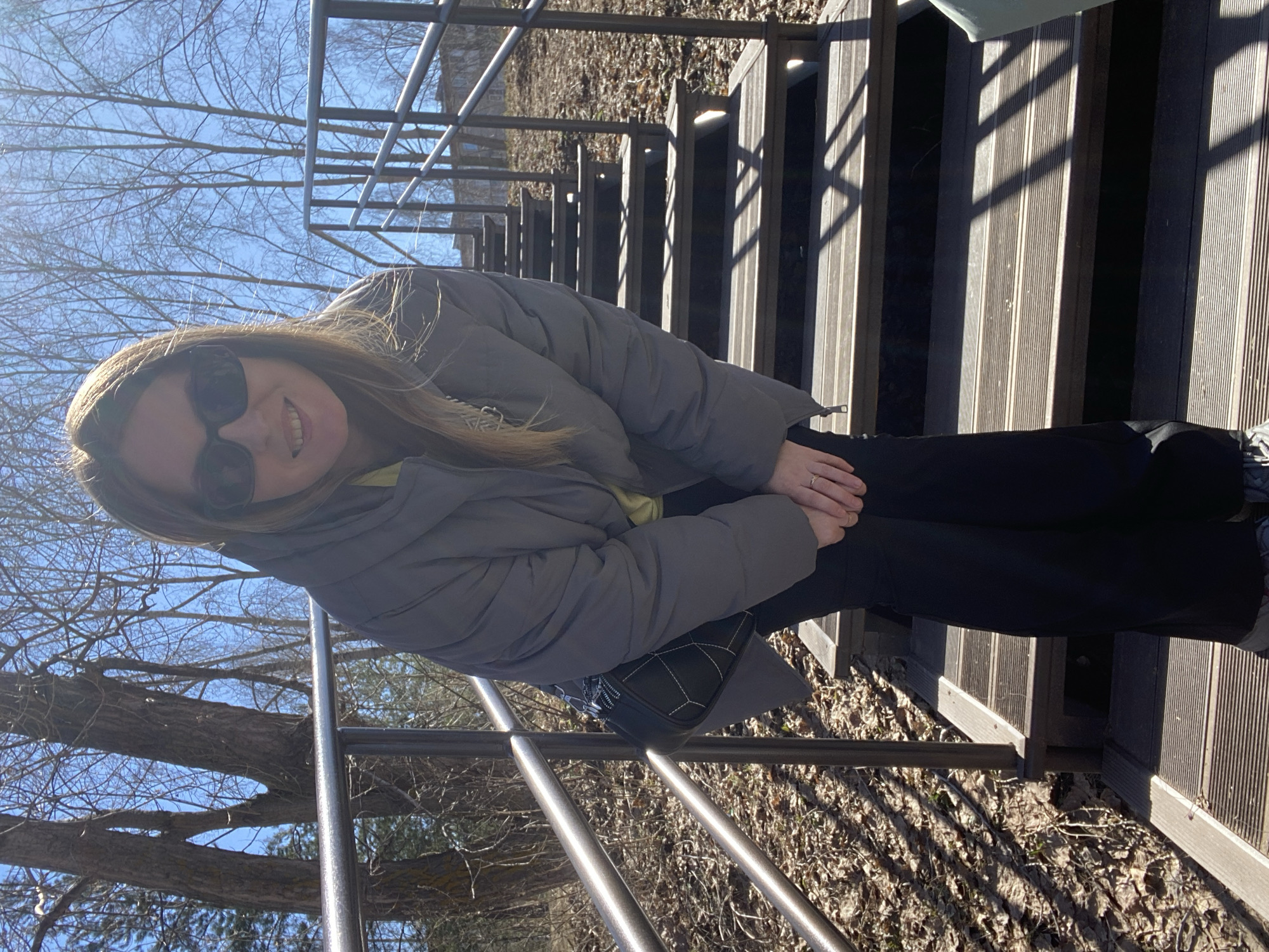A woman wearing a gray puffer jacket, black pants, and sunglasses is sitting on wooden steps with metal railings. She has shoulder-length hair and is smiling. Her hands are clasped in her lap. A black purse is resting against her side. The background includes leafless trees and a clear blue sky. The ground is covered in dry leaves. The location is Chaikovsky, Russia.