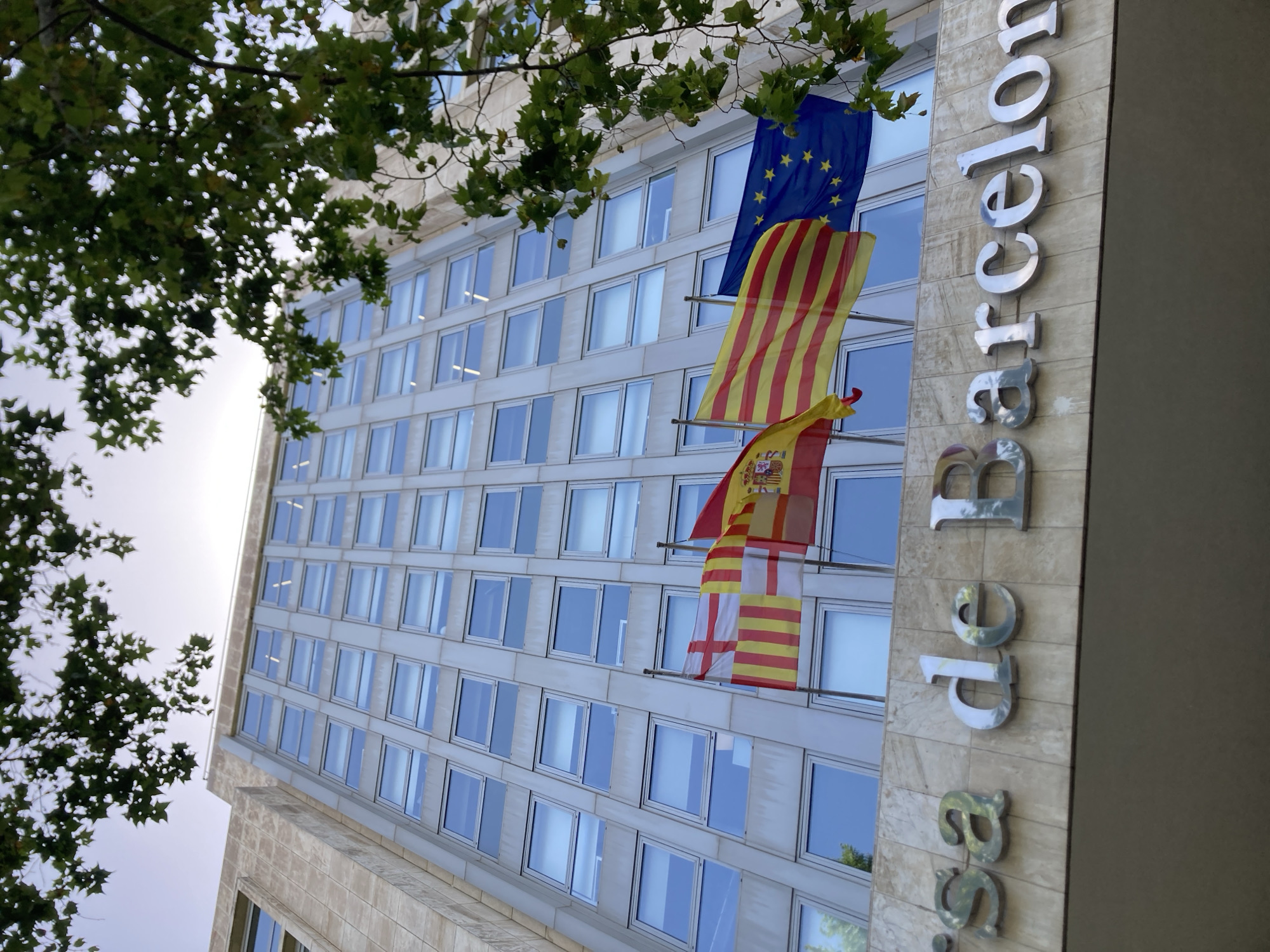 A low-angle, upward shot captures the exterior of a modern, multi-story building in Barcelona, Spain, with large windows and a stone facade. In front of the building, three flags are prominently displayed on poles: the flag of Catalonia, the flag of Spain, and the flag of the European Union. The name "sa de Barcelona" is visible in large, reflective silver lettering above the flags. Green leaves from an overhead tree branch frame the top of the image, partially obscuring the sky. The lighting suggests it is daytime.
