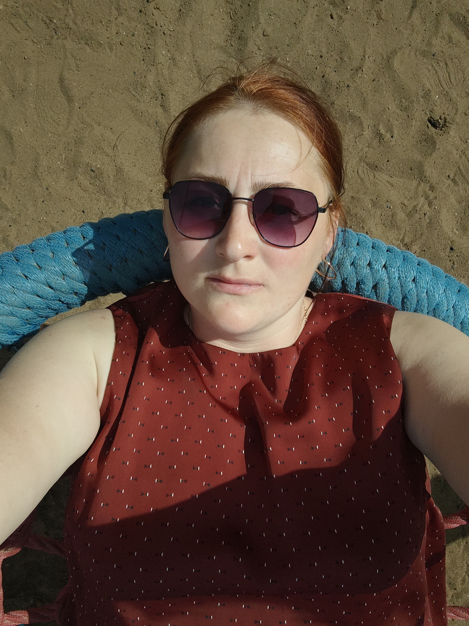 A woman with red hair, wearing sunglasses and a maroon top with a small bow pattern, is lying in what appears to be a hammock. The hammock has blue rope supports. The woman's face is directly facing the camera. The background is sand. The location may be Chaikovsky, Russia.