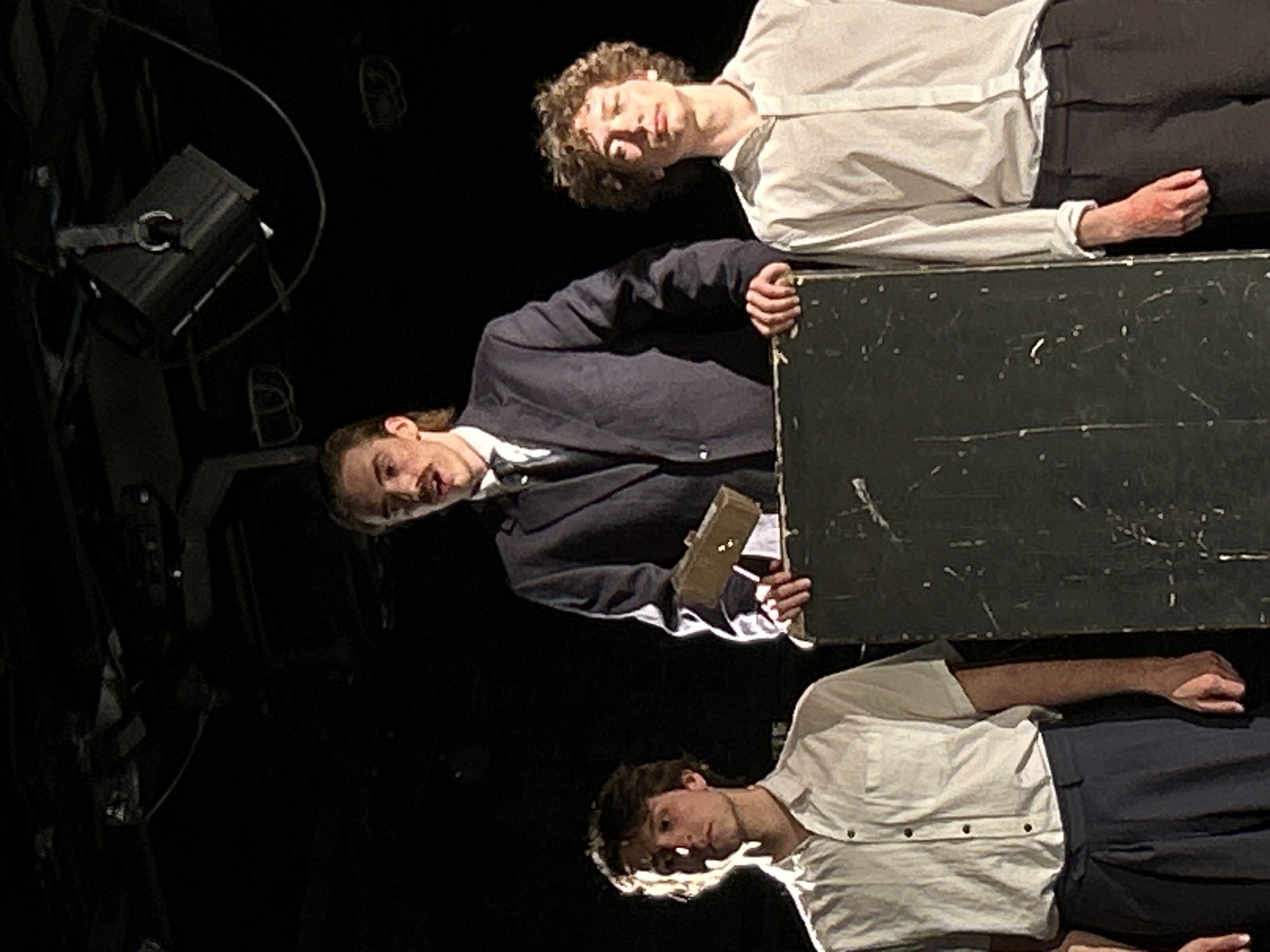 Three individuals are positioned in a theatrical scene. One is standing behind a dark gray rectangular structure, wearing a navy suit jacket, a white collared shirt, and holding a book or small rectangular object. The other two figures are lying down wearing a white collared shirt and dark trousers. Stage lighting fixtures are visible in the background, along with a dark, unlit environment. The setting appears to be indoors, possibly a theater stage. The city location is unknown.