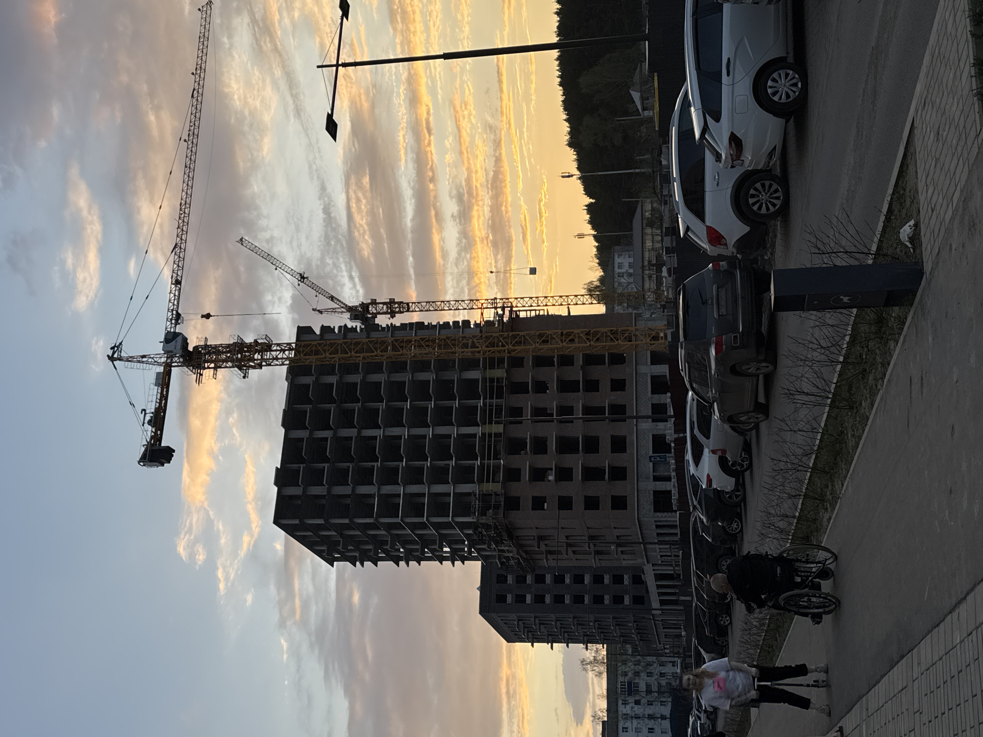 The image captures a construction site in Izhevsk, Russia, at twilight. The partially constructed building, framed by two large cranes, dominates the scene. Two individuals are visible on the sidewalk adjacent to a row of parked cars. One person uses a wheelchair while the other is standing next to them. The environment is a mix of urban construction and residential life. The sky is painted with vibrant sunset colors.