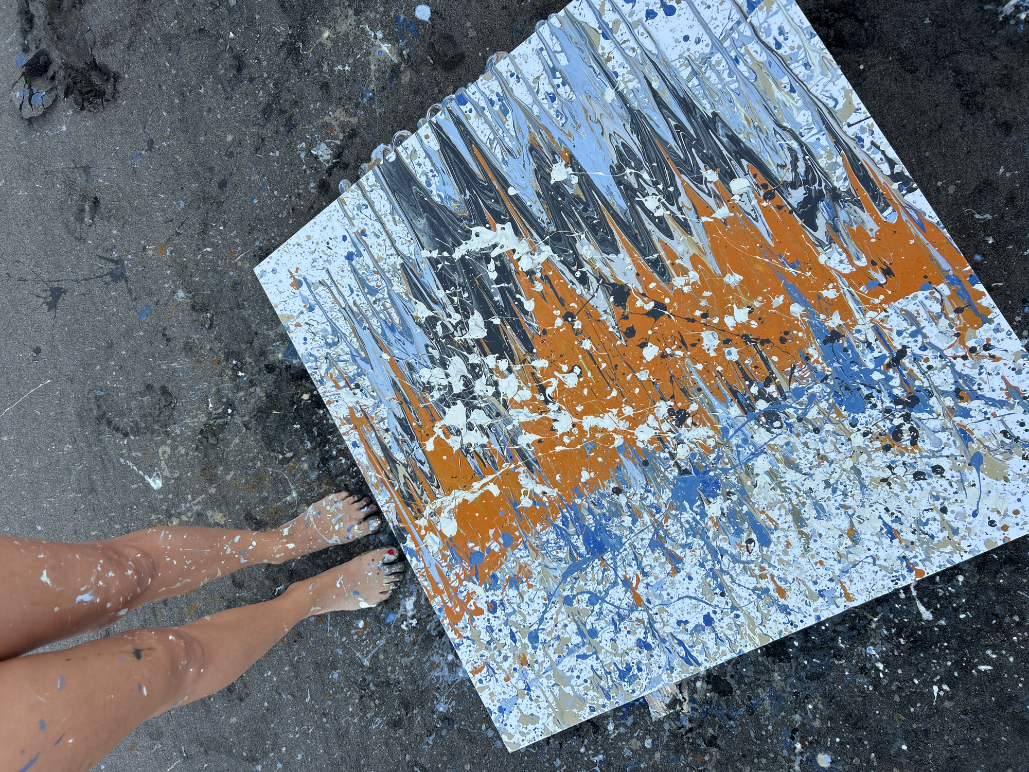 In Tabanan, Indonesia, a person stands barefoot on a paint-splattered ground, their legs also covered in various colored paint splatters. In front of them lies a large, rectangular canvas with a chaotic, abstract design of paint drips and splatters in white, orange, blue, and grey. The ground appears to be asphalt or a similar dark, textured surface, also heavily marked with paint. The scene suggests an outdoor art creation or activity.