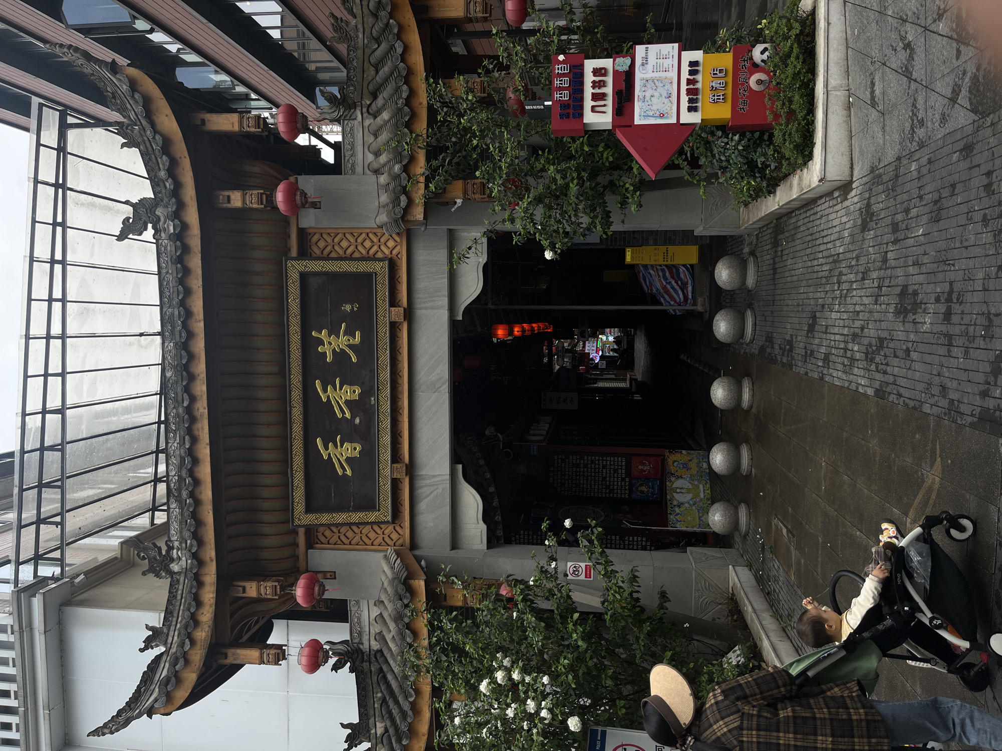 The image shows a street scene in Chenghua District, China, featuring an ornate traditional-style entrance to a shopping area or street. The entrance is marked by a large wooden gate with a curved roof adorned with decorative tiles and dragon-shaped carvings. Two red lanterns hang on either side of the gate, and a sign with Chinese characters is prominently displayed above the entrance. To the right of the entrance are several directional signs with colorful text and graphics, partially obscured by greenery.
The street leading through the entrance is paved with gray stone and lined with bollards. The interior of the street is dimly lit, suggesting it might be a covered area or narrow alleyway. On the right side of the frame, a person is seen pushing a stroller, with a child visible inside. The person is wearing a plaid shirt and a hat. The overall impression is that of a blend of traditional architecture and modern urban life in a Chinese city.