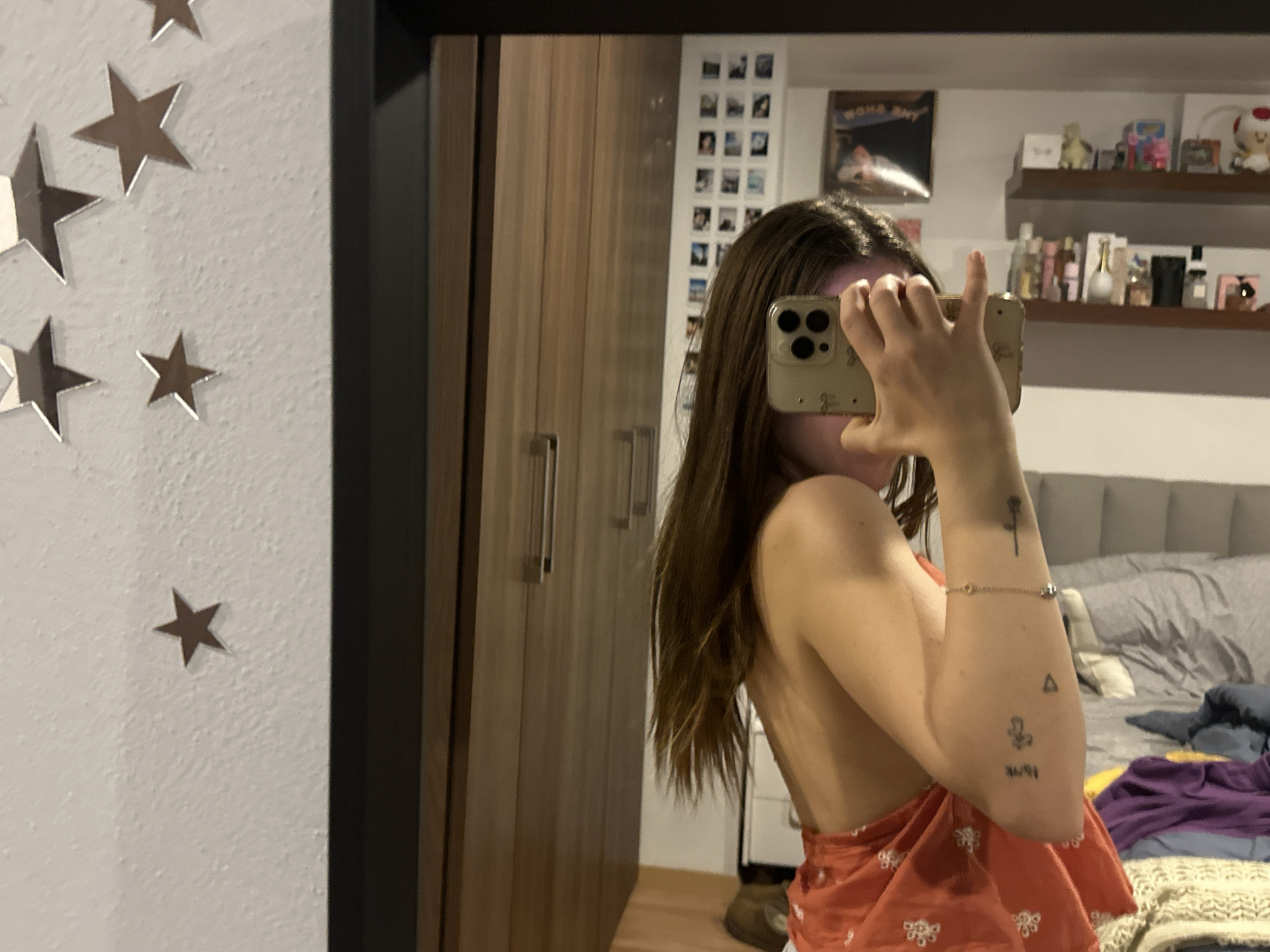 A person stands in front of a mirror, taking a photo with a smartphone. The reflection in the mirror reveals a bedroom with a bed, a shelf with various items, and a wall decorated with small photographs. The mirror frame and a nearby wall feature decorative star-shaped objects. A wardrobe is visible beside the mirror. The person is wearing an orange top. Tattoos are visible on their left arm. The location is a room that is similar to those located in Huixquilucan de Degollado, Mexico.