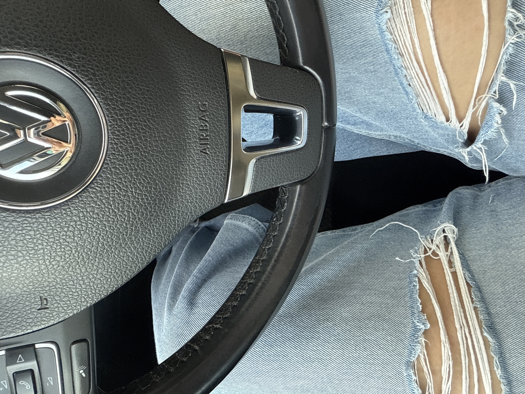 The image shows the interior of a Volkswagen vehicle, specifically focusing on the steering wheel bearing the VW logo and the "AIRBAG" inscription. A person wearing distressed light-wash jeans with prominent rips and frayed edges is positioned behind the steering wheel. The background is dark, possibly indicating the car's interior beyond the visible area. The image does not provide any information about the location in the city of Chaikovsky, Russia.