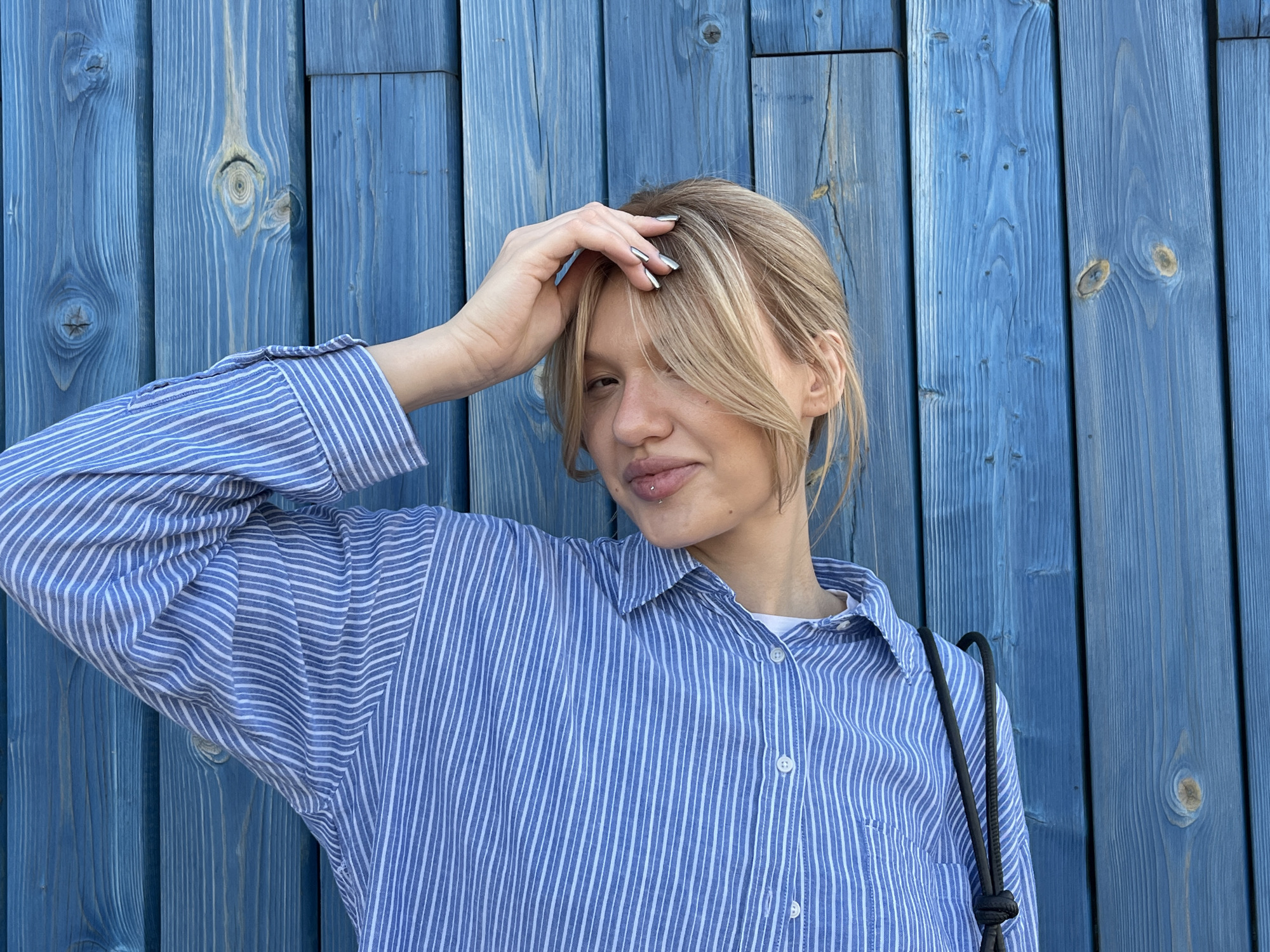 A fair-skinned, blonde woman stands in front of a blue wooden wall. She is wearing a blue and white striped button-down shirt. Her left hand is raised, with her fingers touching her forehead. She has a piercing on her lower lip. A black strap of a bag hangs over her right shoulder. The location is not discernible from the image.