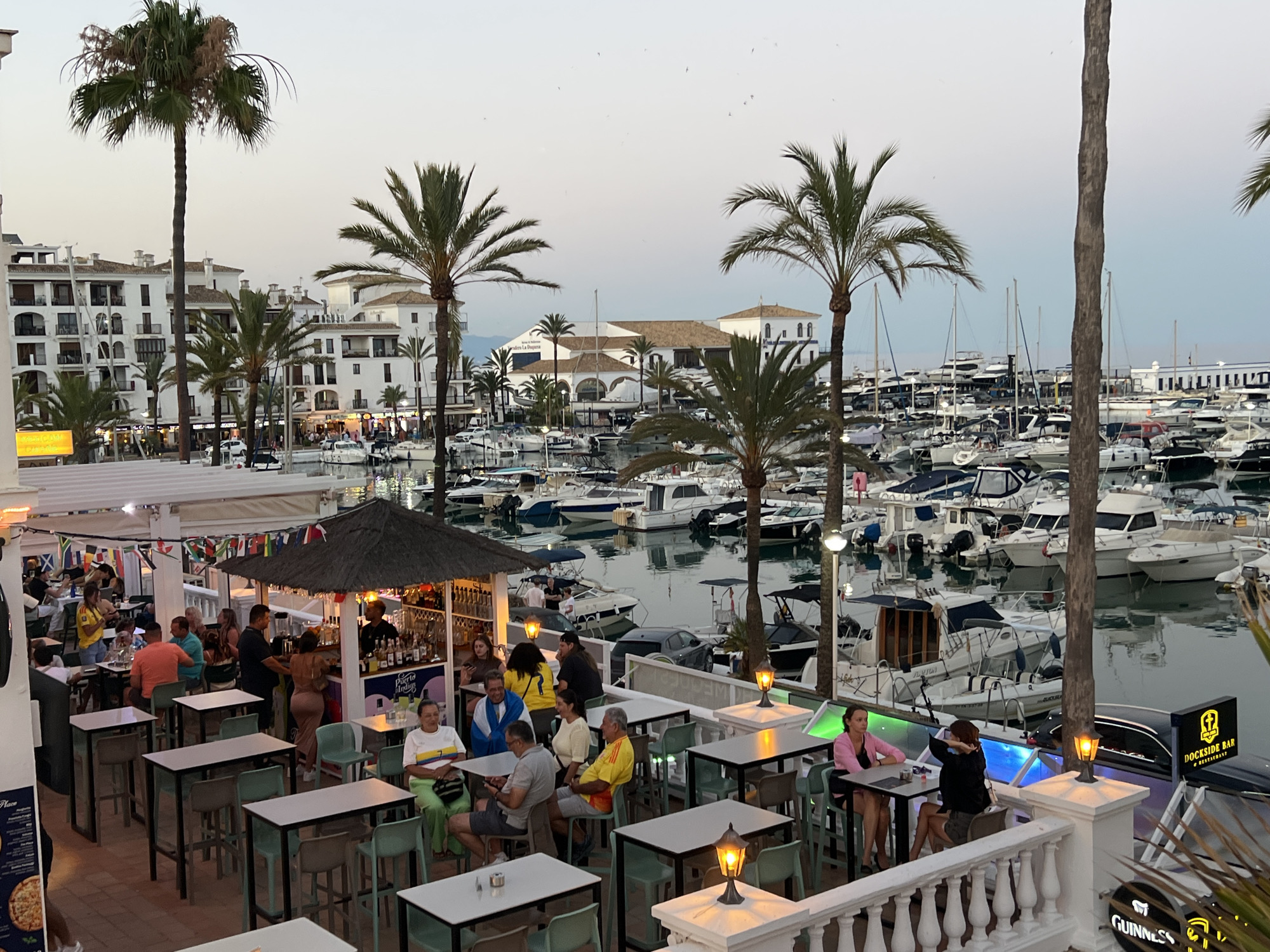 This image depicts a lively outdoor bar and restaurant area in Castillo De La Duquesa, Spain, overlooking a marina filled with numerous boats. The scene is captured at dusk, with artificial lights beginning to illuminate the area and the sky showing soft twilight colors.
In the foreground, several tables and chairs are set up, some occupied by patrons. A bar with a thatched roof is visible, with a bartender serving customers. People are dressed casually, enjoying drinks and food. The bar area is adorned with various flags, suggesting an international clientele or a themed establishment.
Behind the bar, the marina stretches out, packed with a variety of boats, including sailboats and motor yachts. The buildings lining the marina are white and Mediterranean in style, with multiple stories and balconies. Palm trees are scattered throughout the scene, adding to the tropical ambiance.
Further back, more buildings and the silhouettes of distant hills can be seen against the fading light. A black car is parked near the bar area, and a sign for "Dockside Bar & Restaurant" is prominently displayed. Another sign for "Guinness" is also visible. The overall atmosphere appears relaxed and social, indicative of a popular evening spot.