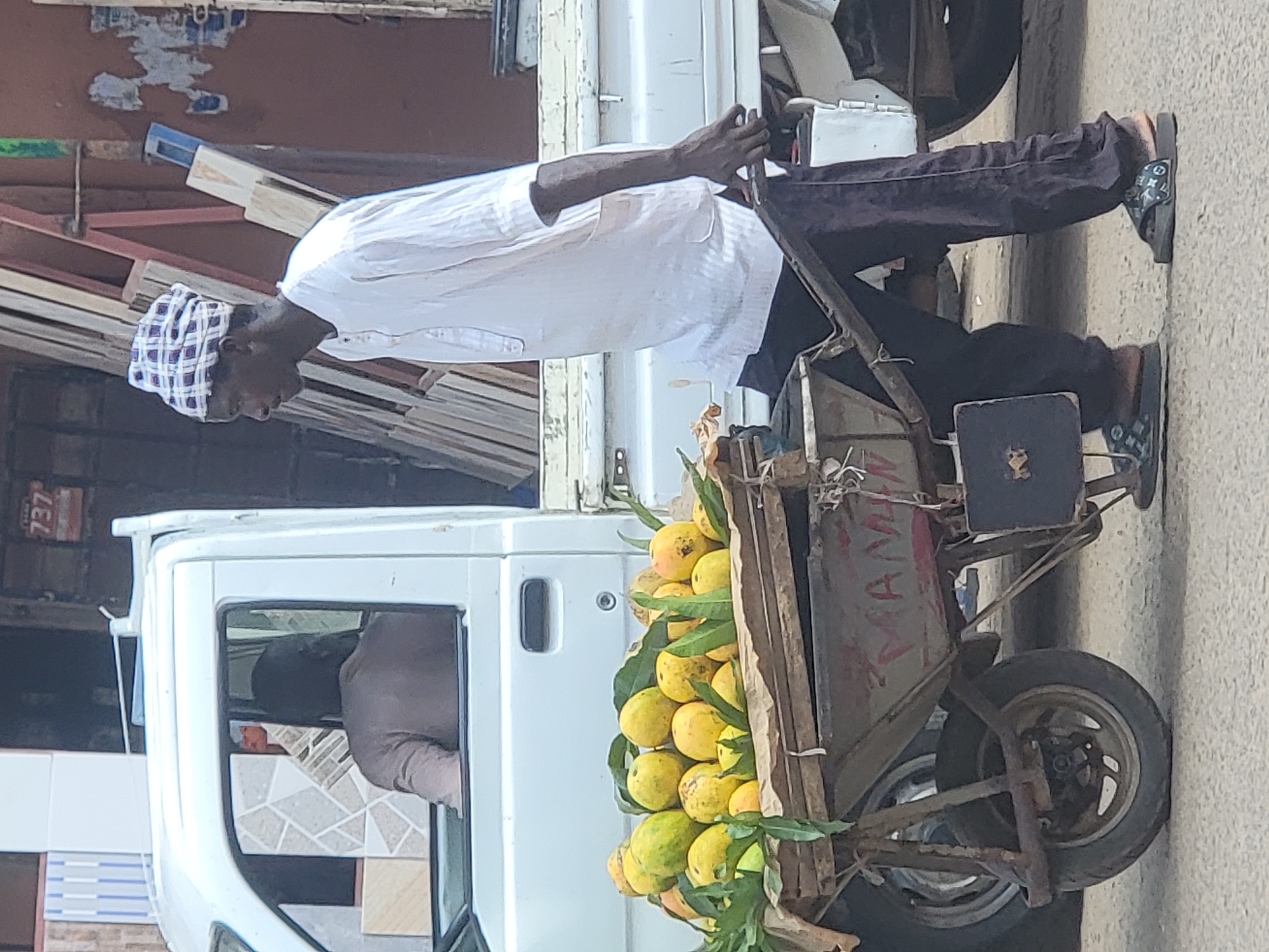 A man in Maiburiji, Nigeria, pushes a handcart laden with mangoes along a street. The man wears a white shirt, dark trousers, patterned slippers, and a patterned cap. The cart is old and rusty. It appears to be constructed of repurposed materials with the word "MAMA" visibly painted on its side. The cart contains a large quantity of yellow mangoes with green leaves. The man is standing on a paved sidewalk. A white truck is beside him. A person is visible in the truck's driver's seat. Tiles are stacked on the truck's flatbed. A building is visible behind the truck.