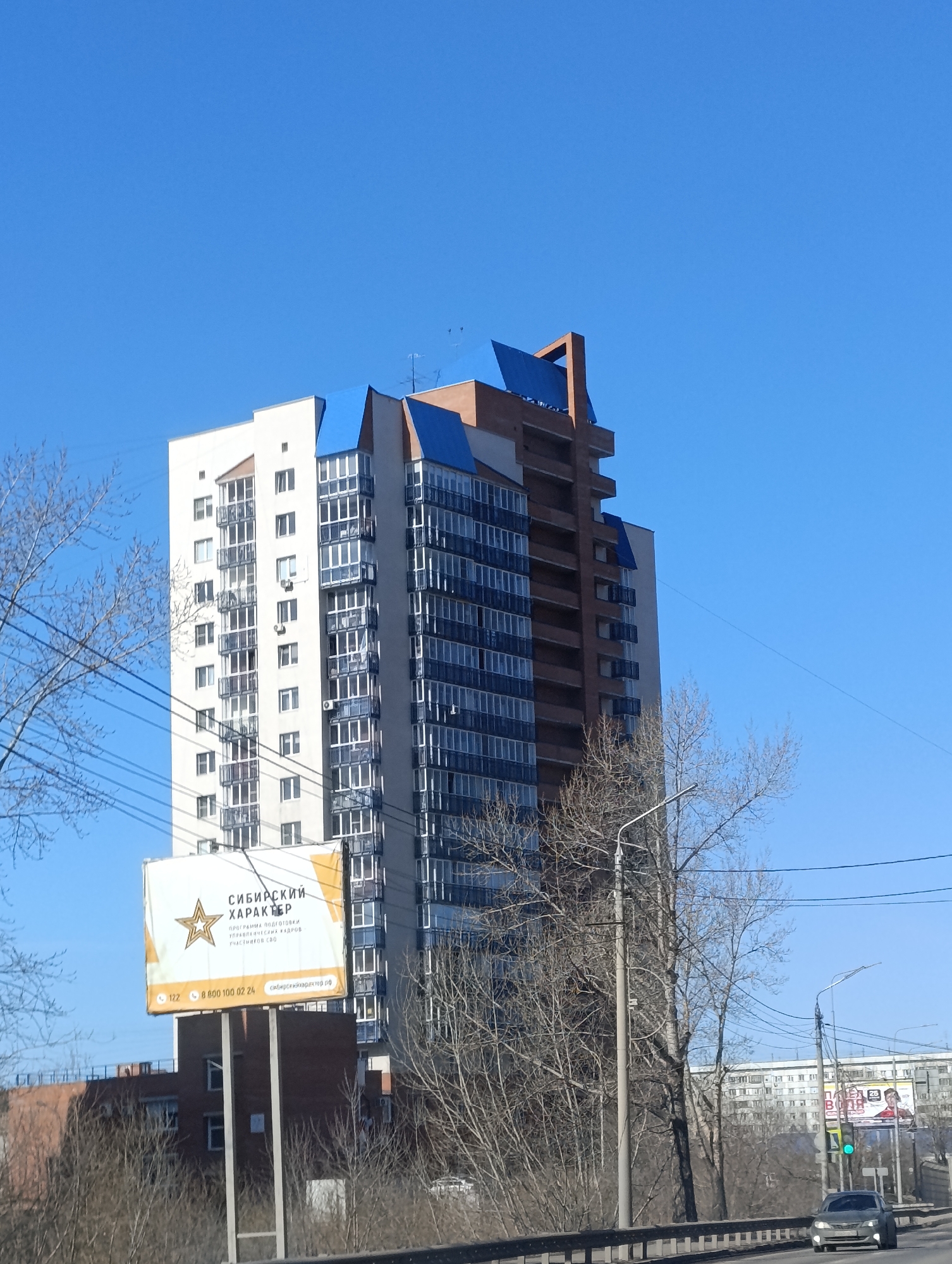 The image depicts a tall residential building in the city of Krasnoyarsk, Russia, notable for its white and brown facade, and blue accents on the upper floors. A billboard with Russian text reading "Siberian Character" is visible in front of the building. Bare trees are in the foreground. A road with a passing car, street lamps, and power lines suggest an urban location. Another residential building is visible further down the road. The sky is clear and blue.