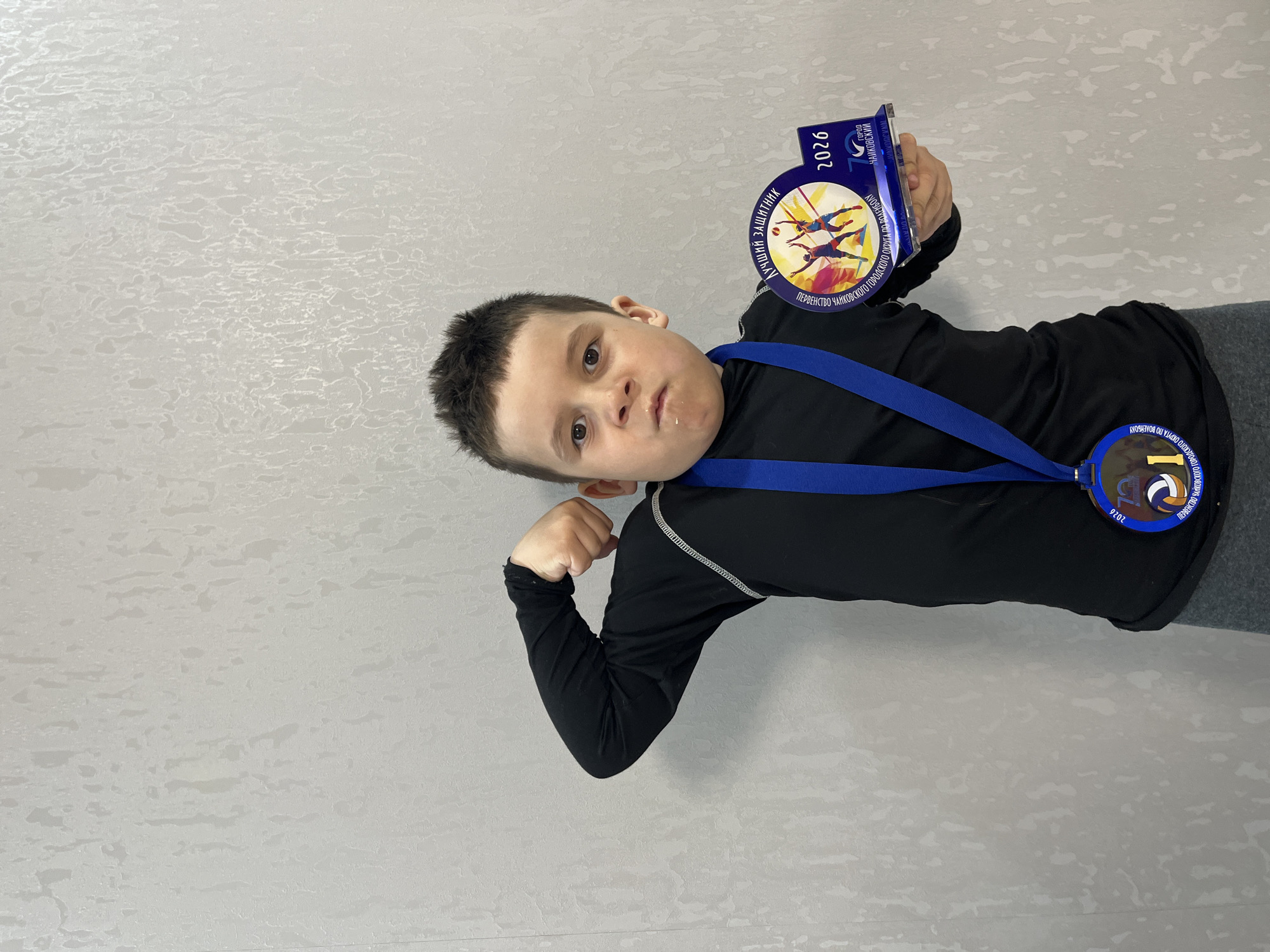 A young boy is standing indoors, wearing a black long-sleeved shirt and grey pants, holding a medal and a commemorative plaque. The medal is attached to a blue ribbon around his neck. The plaque and medal bear the inscription "Первенство Чайковского городского округа по волейболу" and the year "2026", suggesting they are related to a volleyball tournament in the city of Chaikovsky, Russia. He is flexing his left arm and appears to be making a slightly pouting or playful facial expression. The background is a plain, light-colored textured wall.