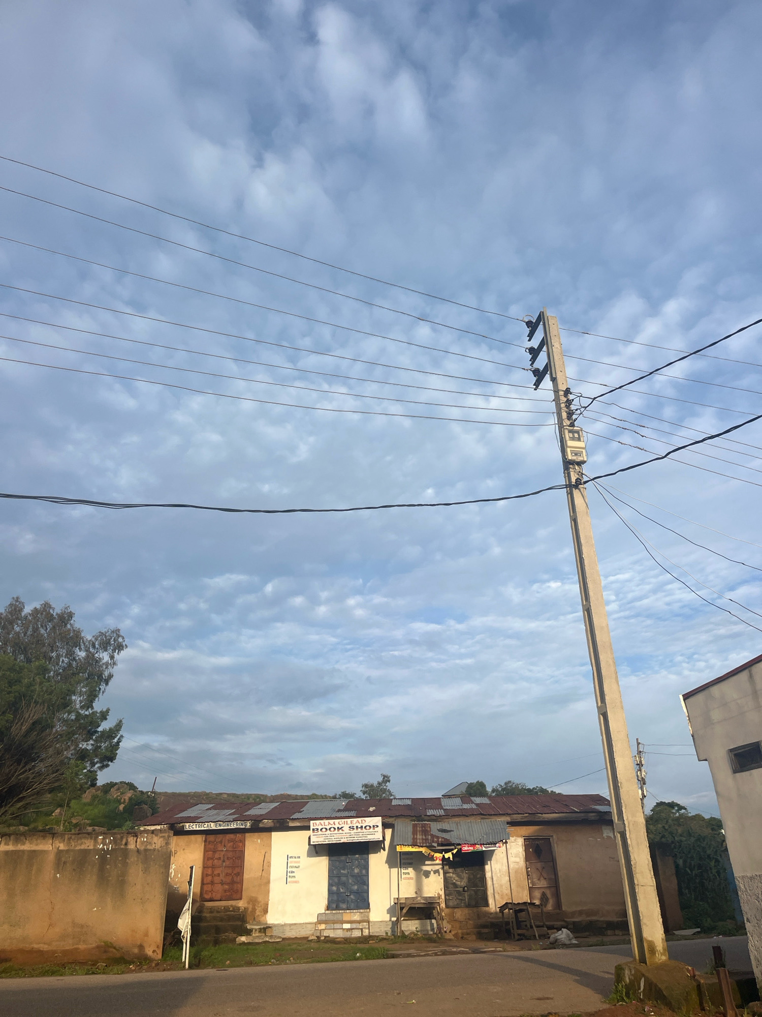 The image shows a street view in Jos, Nigeria, on a cloudy day. The foreground features a paved road and a sidewalk. To the right, a concrete utility pole with electrical wires stretches upwards. Behind the road, a row of modest buildings lines the street. One building prominently displays signs for "ELECTRICAL ENGINEERING" and "BALAH GILEAD BOOK SHOP." Another adjacent building appears to be a shop with a blue metal door. The structures have corrugated metal roofs and weathered walls. A mature tree with sparse foliage stands to the left, next to a stone wall. In the background, a rocky, grassy hillside is visible. The sky is filled with a pattern of scattered white clouds against a blue backdrop.