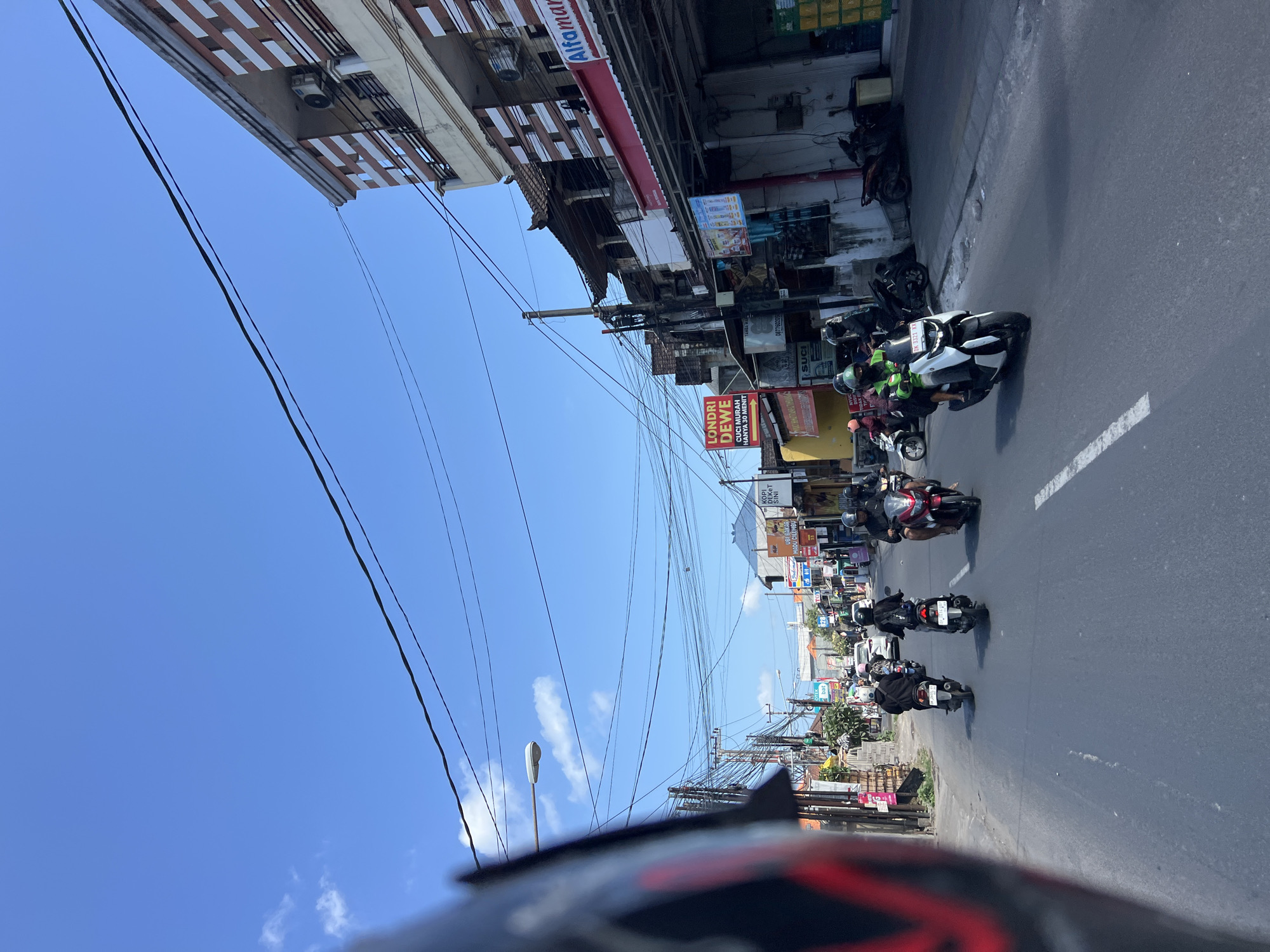 The image depicts a street scene in Denpasar, Indonesia, on a clear day. The foreground shows the left side of a black motorcycle helmet, implying the photo was taken from the perspective of a rider. The road, paved with asphalt and marked with a white line, is populated with several motorbikes and their riders.
In the midground, a convoy of motorbikes is traveling down the street. Most riders are wearing helmets, some are wearing helmets with bright green visors. The buildings lining the street are a mix of commercial establishments and residential structures. One building features an "Alfamart" sign. Another prominent sign reads "LONDRI DEWE CUCI MURAH HANYA 30 MENIT", indicating a laundry service. Overhead, a complex network of power lines and utility poles crisscrosses the sky.
The background reveals more buildings and the clear blue sky with a few scattered white clouds. The overall impression is a bustling urban environment characteristic of Denpasar.