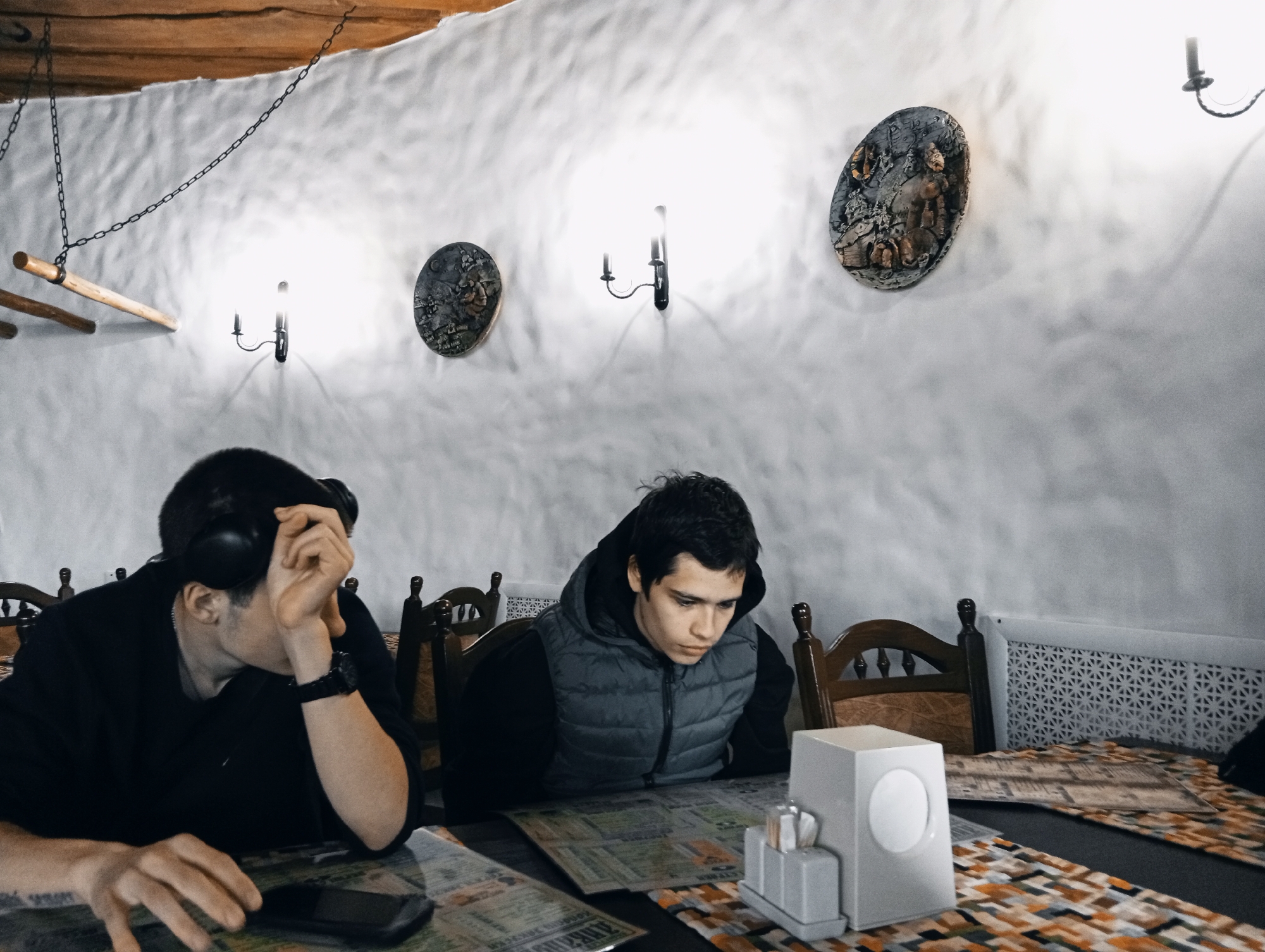 Two young men sit at a wooden table in what appears to be a restaurant. The restaurant is located in Poltava, Ukraine. The man on the left is wearing a black shirt and headphones. He rests his head on his left hand, which is adorned with a black watch. His right hand is near a black smartphone on the table. The man on the right wears a black hooded sweatshirt and a gray quilted vest. Both men are looking at what seems to be a menu or printed material spread across the table.
The table is covered with a patterned placemat in hues of brown, orange, and black. A white dispenser holding napkins and condiments is placed on the table. Behind the men are wooden chairs with decorative backs.
The walls are textured white. Three decorative oval plates are hung on the wall, illuminated by sconce-style lights. A wooden beam structure with hanging chains is visible near the ceiling. A radiator is partially visible on the right side of the image.
