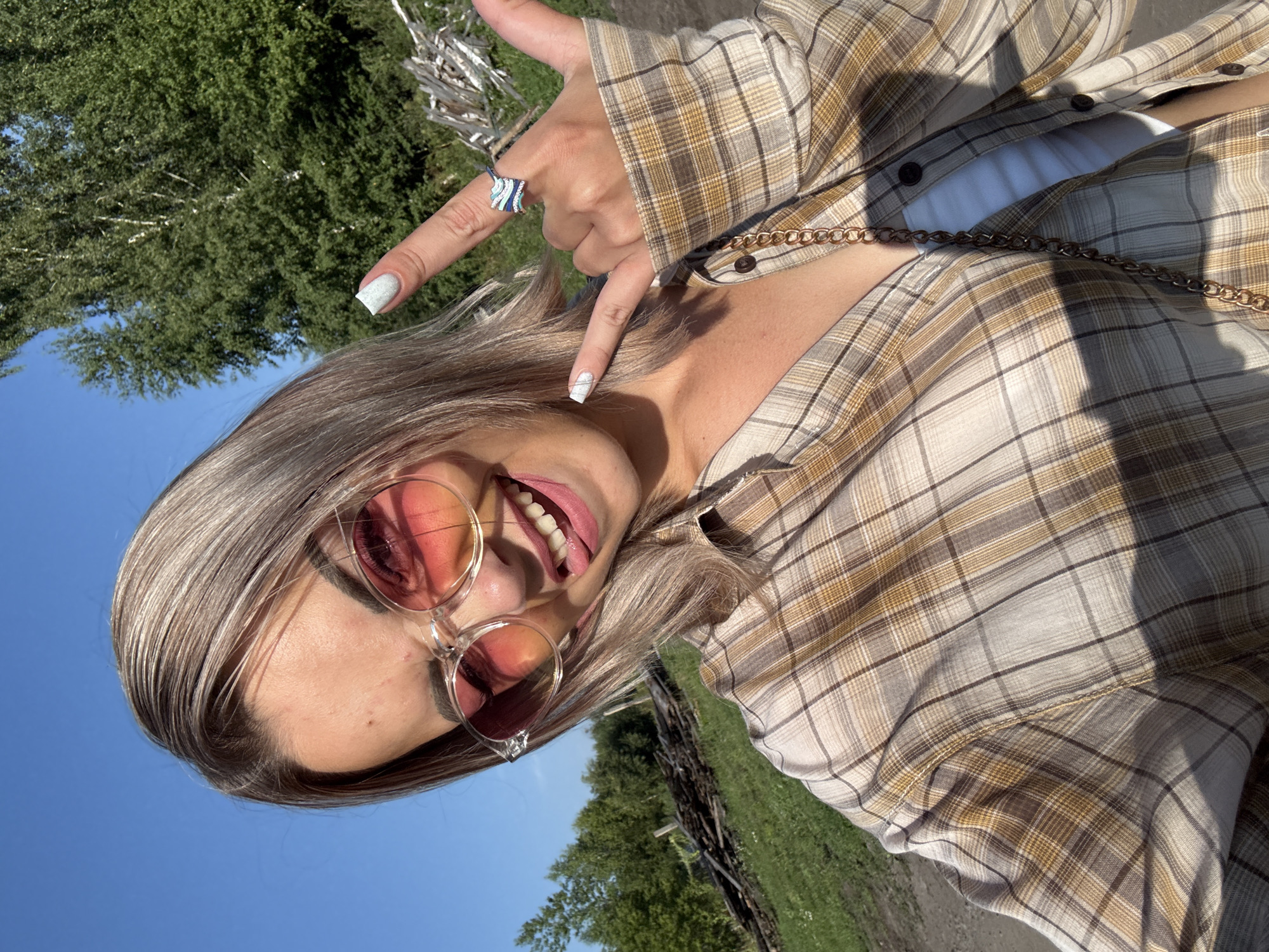 In Krasnogorskoye, Russia, a young woman with blonde hair and pink-tinted sunglasses is captured in a selfie outdoors on a sunny day. She is wearing a plaid shirt over a white tank top and a gold chain. Her right hand is raised, making a rock-on gesture with a ring on her finger. Her left hand is also raised, and she is sticking out her tongue. The background shows greenery, trees, and a clear blue sky.