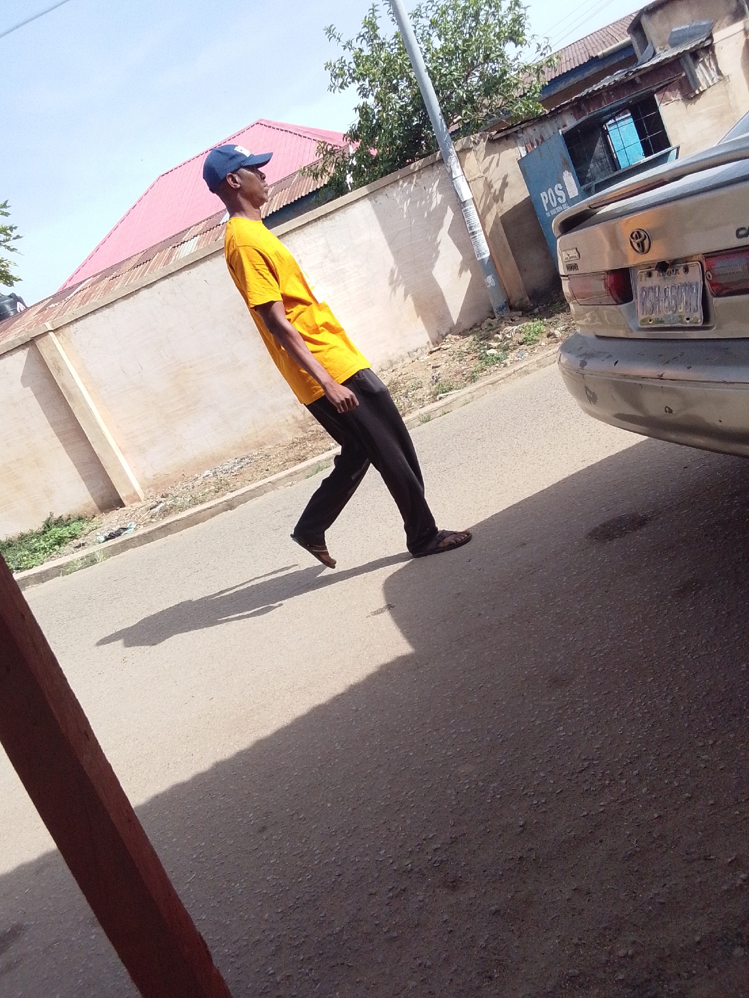 This outdoor image captures a man performing an optical illusion on an asphalt road under a bright, clear sky. The man, dressed in a yellow t-shirt, black pants, and a blue baseball cap, is leaning significantly backward, almost parallel to the ground. His left foot appears to be in contact with the ground, while his right foot is lifted, creating the impression that he is defying gravity. He is wearing brown sandals. A long, distinct shadow of the man is cast on the road, indicating strong sunlight, likely around midday or early afternoon.
In the background, a light-colored concrete wall stretches across the scene. Behind the wall, a building with a prominent red-tiled roof is visible. To the right of the man, the rear of a silver Toyota car is parked, partially obscuring a blue structure. The car's license plate is visible, reading "REDIA" at the top and "BGH-860KUY" below. The blue structure behind the car has "POS" written on it in large white letters, with smaller text underneath that appears to say "FOR YOUR NEXT BILL." A gray utility pole stands near the car, with some faint text or notices attached to it. Green foliage is scattered throughout the background, both near the buildings and around the utility pole. The ground is an asphalt road with a dirt strip and some sparse greenery along the curb. The setting appears to be a sunlit street in a residential or mixed-use area, consistent with the provided context of Jos, Nigeria.