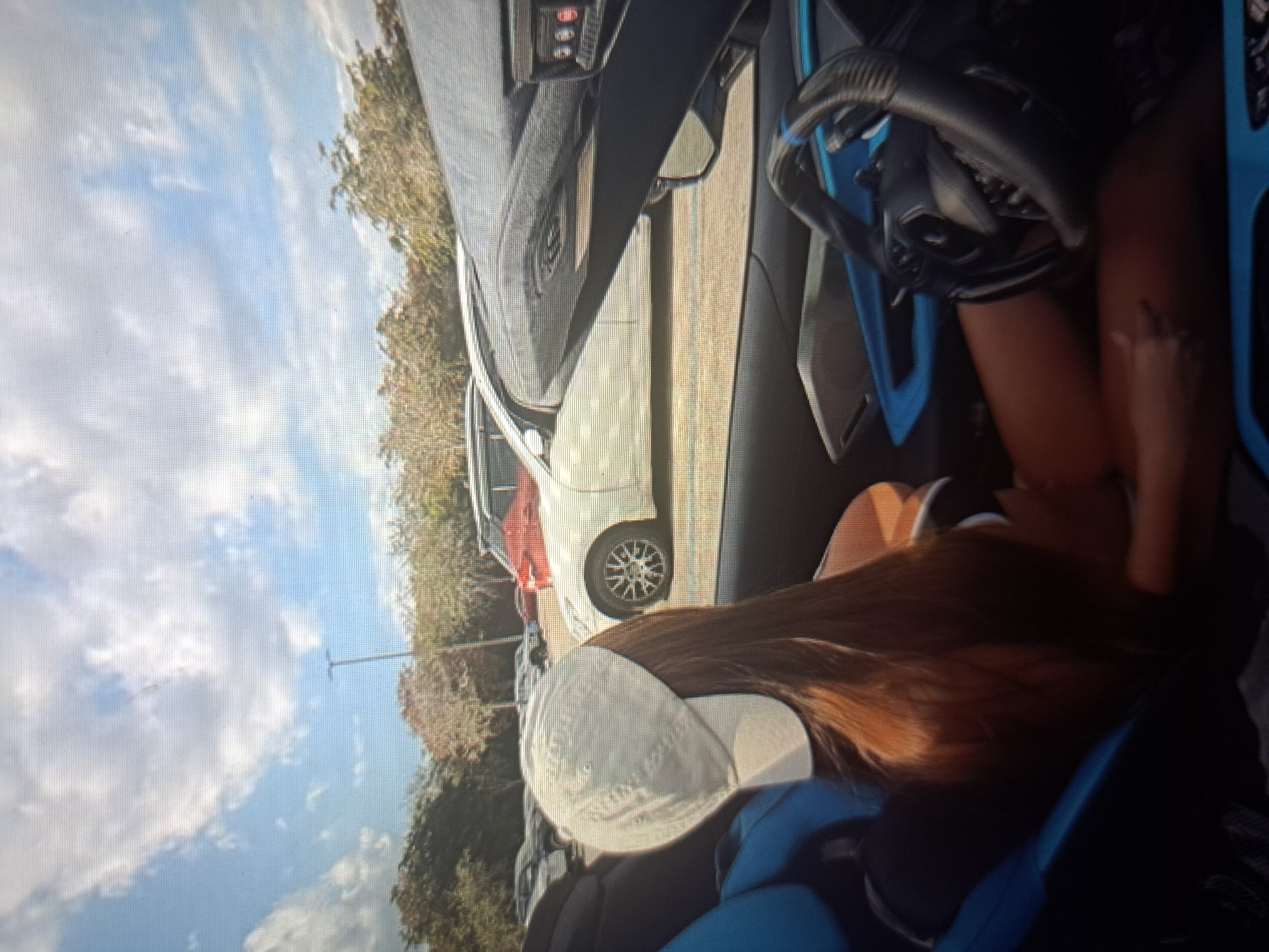 The image shows a person sitting in the driver's seat of a car parked on a street. The person has long brown hair and is wearing a white baseball cap. The car is parked on a street with other cars visible in the background. There are trees and a blue sky with clouds in the background. The location is likely Yerevan, Armenia, but there is no specific landmark visible in the image to confirm this. The person is facing forward, possibly looking at the road ahead. The steering wheel and dashboard are visible inside the car.