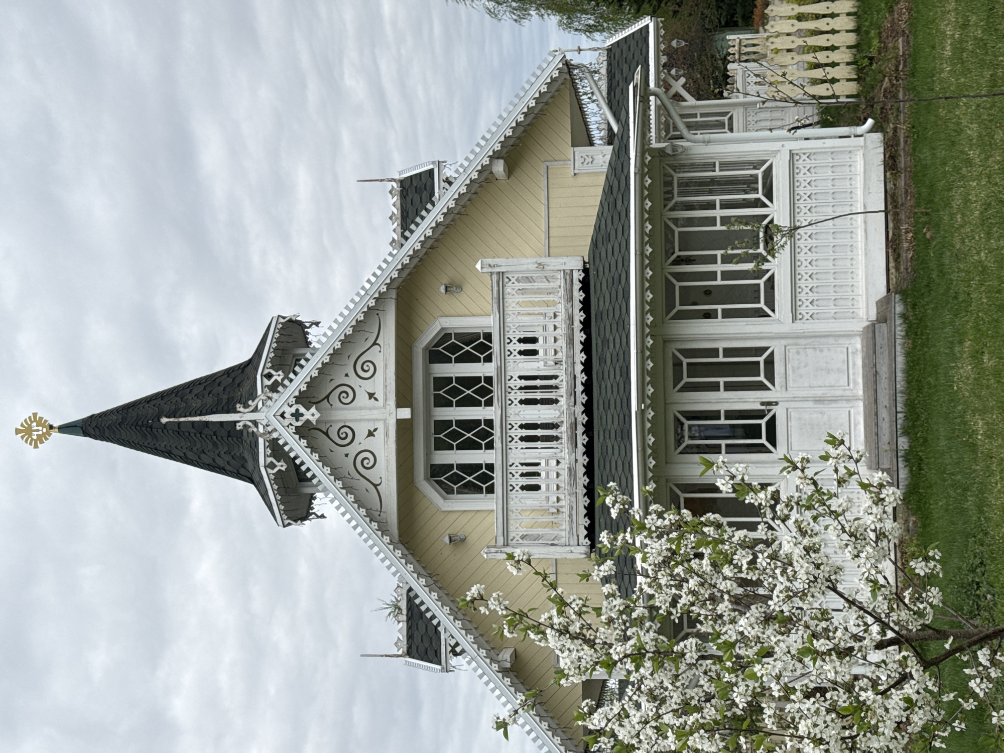 The image shows a detailed view of a wooden house with elaborate ornamentation, located in Sapronovo, Russia. The house is predominantly painted in a pale yellow color, with white decorative trim and a dark, possibly black, roof. The facade features a pointed turret or tower element topped with a golden ornament. A prominent balcony area is visible with white railings. The house has large windows with decorative grid patterns. In the foreground, a tree with white blossoms is in full bloom, partially obscuring the lower portion of the house. Green grass is visible on the right, suggesting a yard or garden. The sky is overcast. The image composition is vertically oriented.