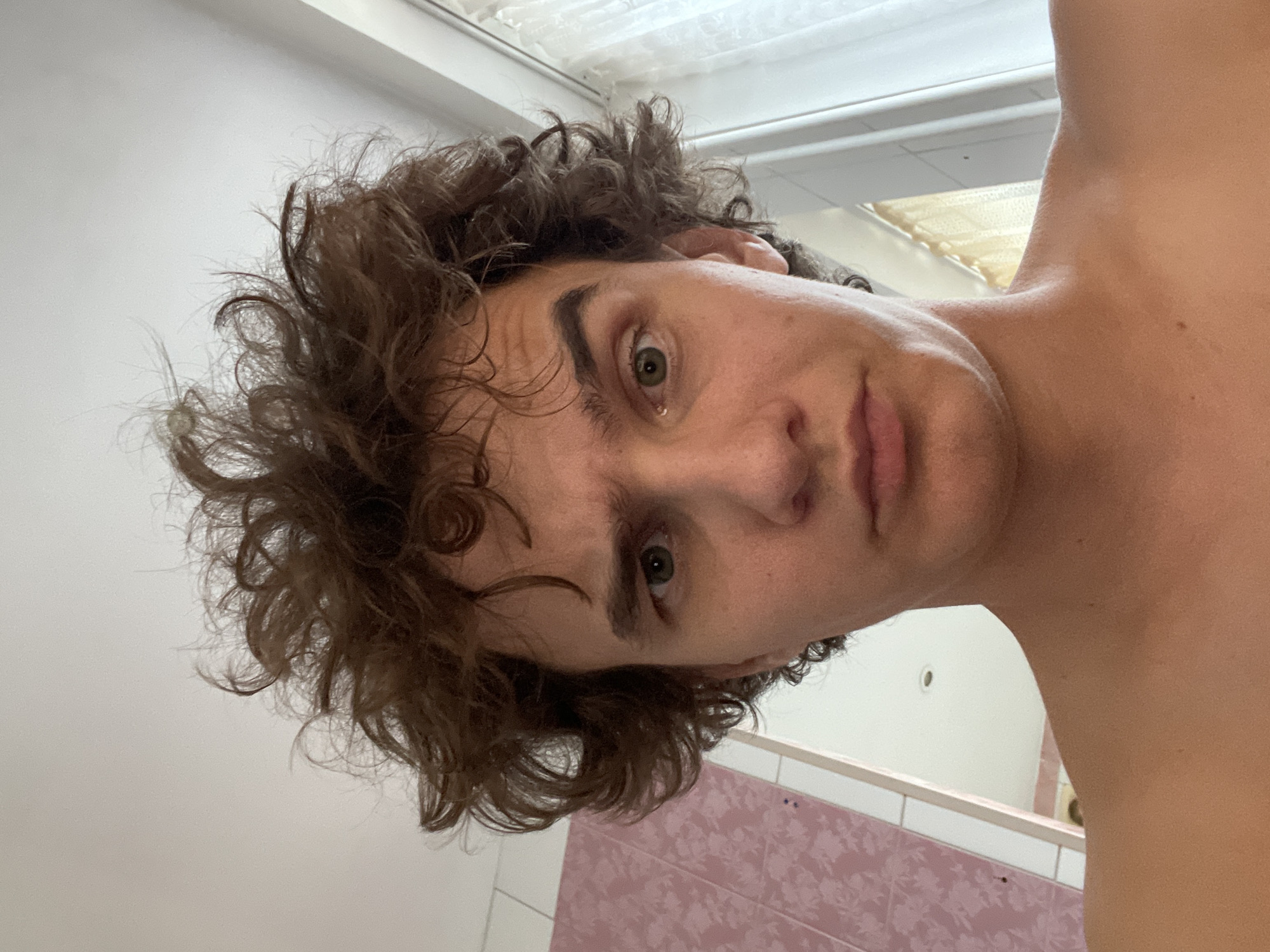 The media file is a selfie of a young person with curly brown hair, taken in portrait orientation and rotated 90 degrees counterclockwise. The subject's face is partially visible, with a green eye looking towards the camera. Their skin appears fair and smooth. The background is out of focus, but suggests an indoor setting. Elements in the background include a white ceiling with fluorescent lighting, a pink tiled wall with a floral pattern, and a white wall. The lighting appears to be artificial. The subject's expression is neutral. There is no visible text in the image. The context provided is Krasnoyarsk, Russia.