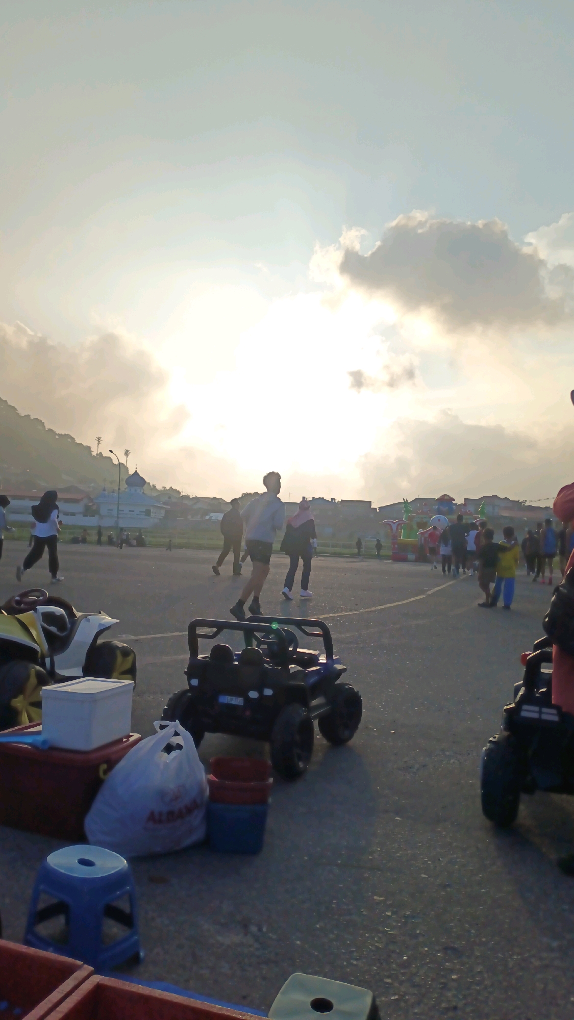 The image depicts an outdoor scene in Padang Panjang, Indonesia, likely at a recreational area or park, with a view of buildings and hills in the background. The sky is bright with soft clouds, suggesting either sunrise or sunset, with the sun obscured. In the foreground, there are several children's ride-on vehicles, including a black toy jeep. To the left, a white cooler sits on a red storage box, accompanied by a white plastic bag with the word "ALBANA" visible. A small blue stool is also present.
In the mid-ground, a group of people are walking and running on an asphalt surface. Some are wearing casual attire, including a woman in a headscarf. Further back, there are colorful inflatable structures, possibly bouncy castles, and more people gathered. A prominent blue-domed building, resembling a mosque, is visible in the distance, situated among the houses on the hillside. The overall atmosphere suggests a lively community gathering or event.