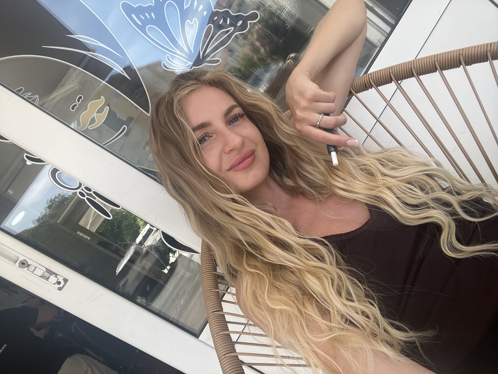 A blonde-haired woman in a brown tank top sits in a wicker chair, holding a vaping device in her right hand. She is looking directly at the camera with a slight smile. The background features a window with a white frame and decorative butterfly design. Reflected in the window are buildings and greenery, suggesting an urban setting. The location is Budva, Montenegro.