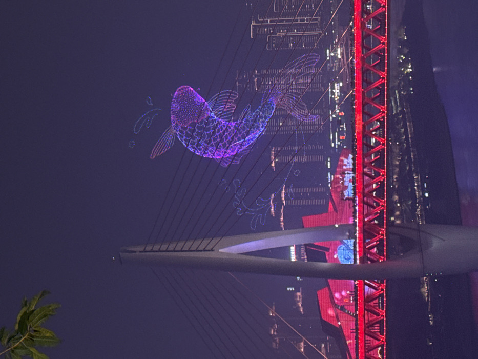 The image displays a night scene in Yuzhong District, Chongqing, China, featuring the Dongshuimen Bridge and a drone light show. The sky is dark, against which a large, illuminated drone formation, resembling a purple and pink koi fish with stylized water splashes, is prominently featured in the upper right. A smaller, similar fish shape is visible to the left of the main formation. A tree branch with green leaves partially occupies the upper left corner.
The Dongshuimen Bridge spans across the midground, characterized by a tall, light-colored A-frame pylon on the left, from which multiple dark cables extend diagonally across the frame. The bridge's lower truss structure is brightly illuminated in red, with a line of smaller white lights along its upper edge. Numerous indistinct figures, consistent with pedestrians, are visible on the bridge deck.
Below the bridge, a dark body of water reflects ambient city lights. In the background, a dense urban skyline with multiple high-rise buildings is visible across the river, many illuminated, including a large building complex with reddish lighting behind the bridge's pylon.