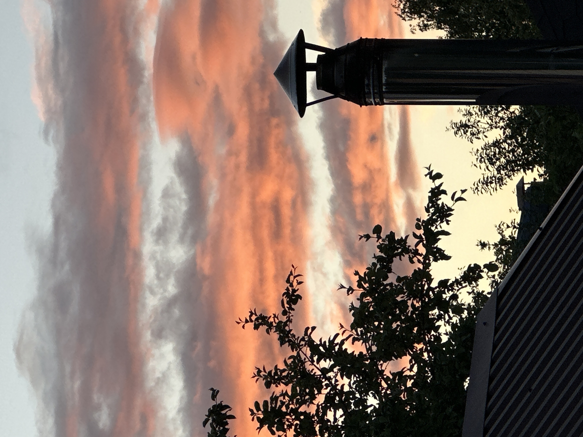 The image, rotated counter-clockwise, shows a sunset sky with striated orange and grey clouds above trees and rooftops. The sky is the prominent feature, filling most of the frame. Silhouetted against the sky is a dark metal chimney with a conical cap on a nearby building. In the foreground are tree branches with dark leaves. A portion of a rooftop with dark diagonal lines is visible in the lower right corner. The exact location within Nizhny Novgorod, Russia, cannot be determined from this image alone. No people are visible.