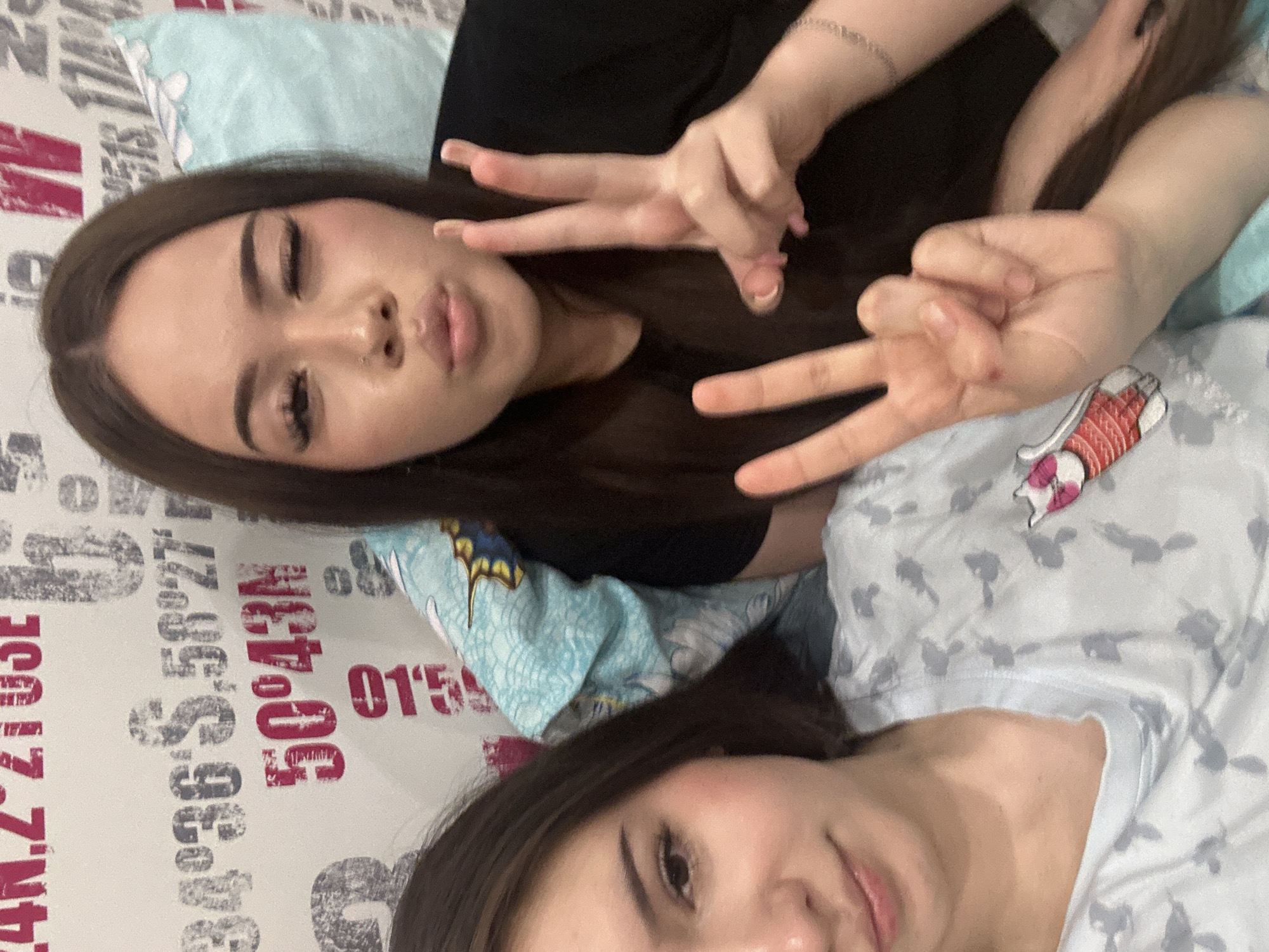 Two young women are depicted in a close-up, selfie-style photograph. The woman on the right is winking and making a peace sign with her right hand, while her left hand is partially visible in the lower left corner, also in a peace sign gesture. She is wearing a black t-shirt and a thin silver bracelet on her left wrist. The woman on the left is looking at the camera with a slight smile. She is wearing a light blue t-shirt with a repeating pattern of grey bird-like figures and a pink and white bird appliqué on the left chest. The background consists of a white wall with large, stylized lettering, including "34°36'S, 58°27'" and "50°43'N" in pink and grey. A light blue pillow with a floral pattern is visible behind the woman on the right. The image appears to be taken indoors, likely in a bedroom. There is no information to ascertain the location within Naberezhnye Chelny, Russia.