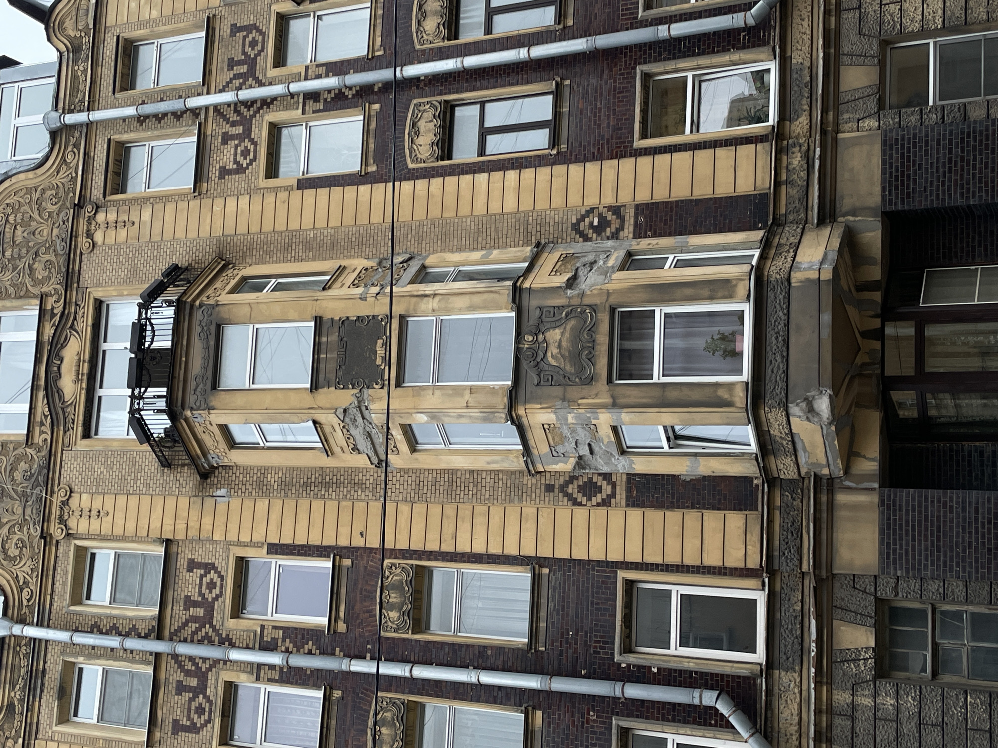 The image captures the facade of a historic building in Saint Petersburg, Russia. The building features a mix of brick and stone construction with detailed ornamentation around the windows and along the vertical edges of the building. The color palette is dominated by browns, tans, and grays. A notable architectural feature is a projecting bay window structure, displaying deteriorated areas. Several pipes run vertically along the building's exterior. A small wrought-iron balcony is visible on the left side. The windows are mostly closed, and the sky visible through them is overcast.