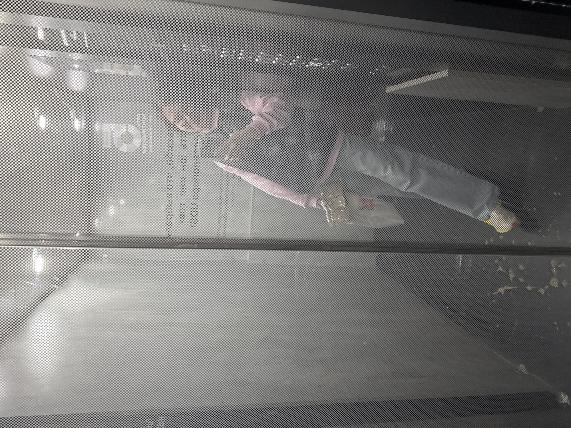 The image captures an indoor scene viewed through a fine, perforated screen, creating a distinct checkered overlay across the entire frame. In the upper right quadrant, a doll or mannequin is prominently featured, dressed in a pink hooded top, light blue jeans, and yellow footwear. It is in a bent-over posture, holding a clear plastic bag or object in its hands. Behind the doll, a white shelf is visible.
The background shows reflections of artificial lighting, possibly fluorescent fixtures. Several pieces of text are visible, either reflected or printed on surfaces behind the screen. In the upper left, large letters read "TO," with partially inverted Cyrillic text above it appearing to say "ХОЛОДИЛЬНИК В ОГОРОЖЕННОЙ" (Refrigerator in an enclosed area). Below "TO," the words "000 PRO" or "300 PRO" and "SENSOR PRO" are discernable. Faint numbers such as "788" are also present near the doll's head. The bottom right area shows some smudged or painted white marks.
The scene appears to be a static display, possibly within a shop, museum, or an artistic installation, with the doll arranged as part of the exhibit. The artificial lighting indicates an indoor setting. The textural interference from the perforated screen is a key visual element of the photograph. The context places this scene in Izhevsk, Russia.