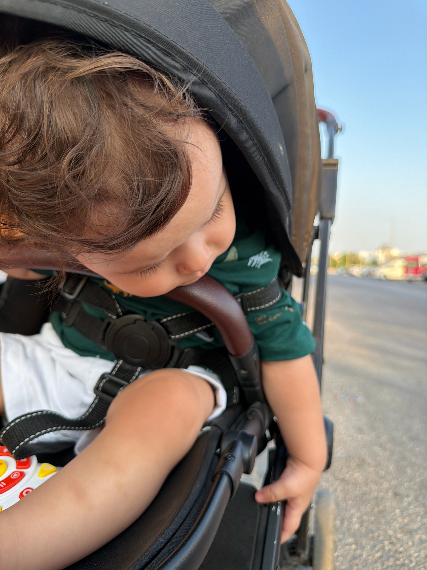 A close-up, high-angle shot shows a toddler sitting in a stroller. The child is wearing a dark green t-shirt and white shorts, with a black stroller harness secured around them. Their hair is brown and slightly messy, and they are looking downwards. The stroller's canopy is a dark color, and the background shows a blurred outdoor scene with a road and buildings, suggesting a street in Sharm El Sheikh, Egypt. The lighting indicates it is daytime.