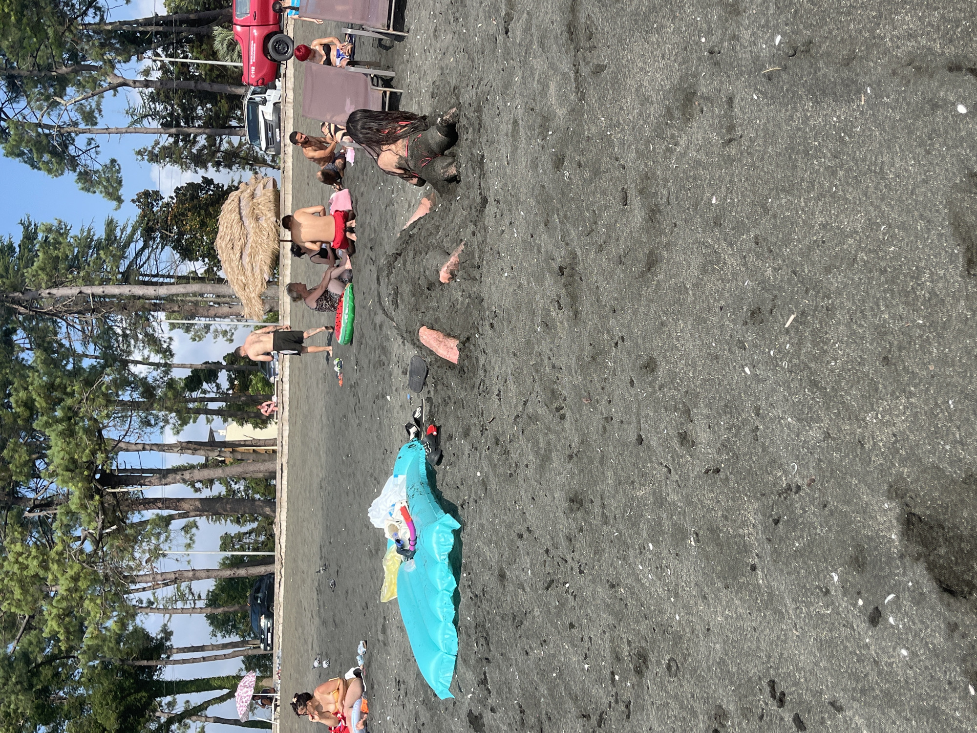 The image depicts a beach scene in Ureki, Georgia, a location known for its magnetic black sand. In the foreground, a person is partially buried in the sand, with their legs and torso visible. Another person is seated in the sand nearby. The beach is populated with other individuals engaged in various activities, including sunbathing on lounge chairs, sitting on the sand, and walking. An inflatable pool float lies on the sand in the middle ground. The background features a line of tall pine trees, a road with parked vehicles, including a red truck, and some buildings. The sky is clear and blue. The overall atmosphere suggests a typical summer day at a seaside resort.