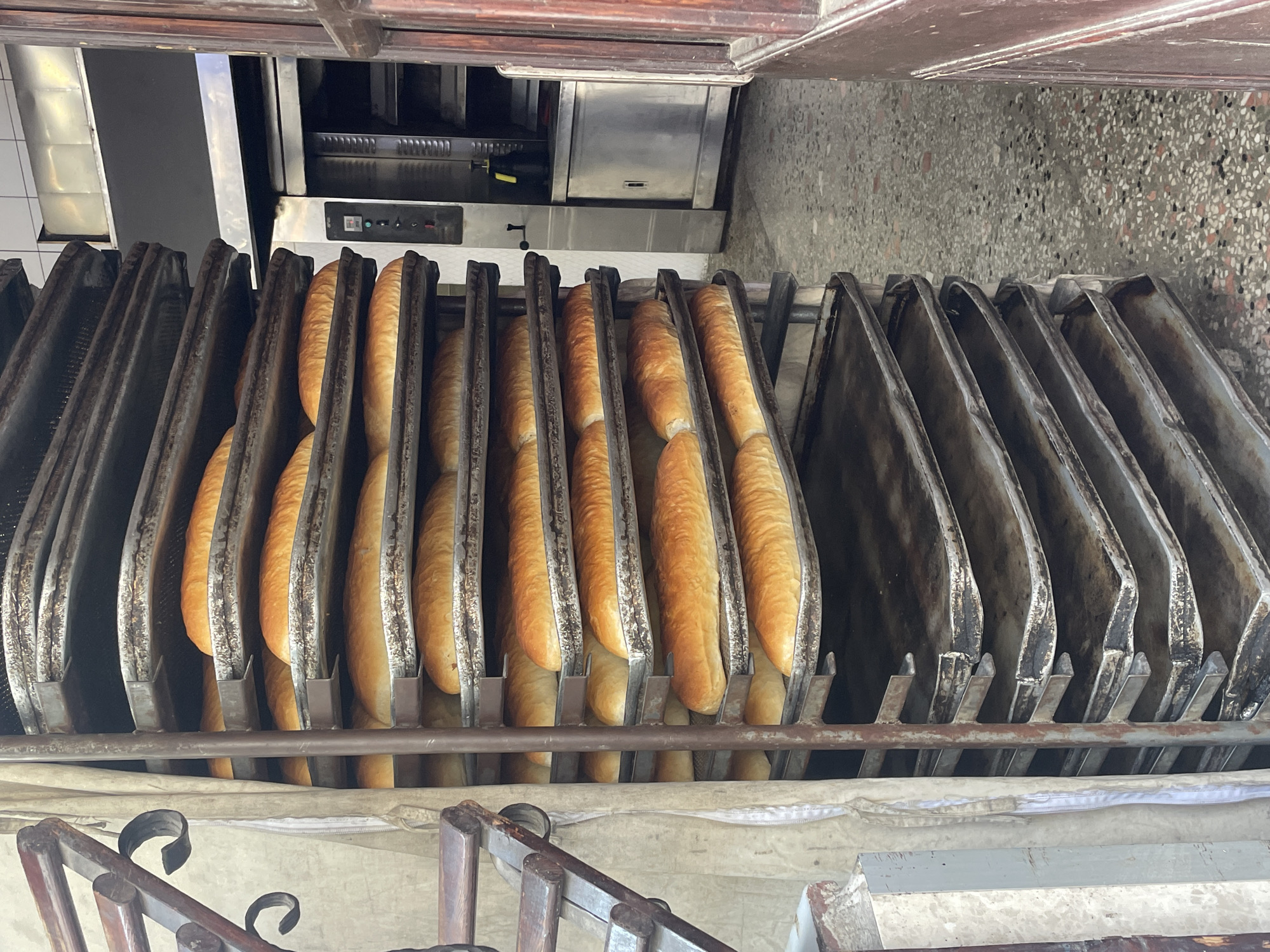 The image depicts a rack filled with freshly baked loaves of bread, stacked on metal trays. The rack is positioned next to a rustic-looking oven, with part of a brown wooden door visible on the right side of the frame. The floor is covered in a speckled terrazzo pattern. The bread appears golden brown and ready for consumption. The scene suggests a bakery or food establishment, possibly located in Kadıköy, Turkey, given the stylistic elements and common bakery scenes found in the region. There are no people visible in the image. The lighting is natural, indicating it could be daytime. The overall impression is one of a bustling, artisanal food production environment.