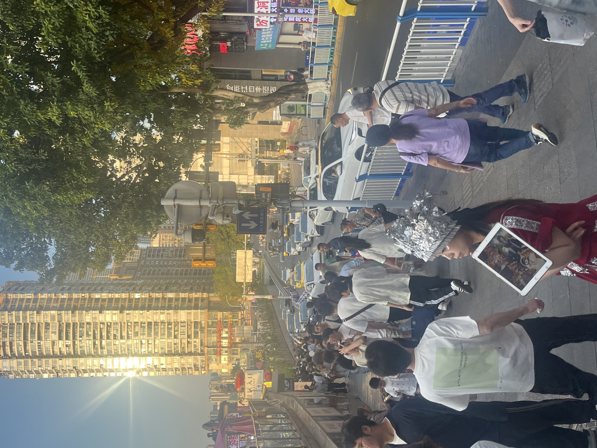 The image captures an outdoor scene in Yuzhong District, China. In the foreground, pedestrians are walking along a sidewalk next to a street with traffic. A person is wearing a reflective head covering and holding an iPad, facing the camera. The background features tall buildings reflecting sunlight, trees, and street signs in Chinese characters. The sky is clear and blue. A road with numerous vehicles is visible, suggesting a busy urban area. The composition suggests an evening scene with the sun setting.