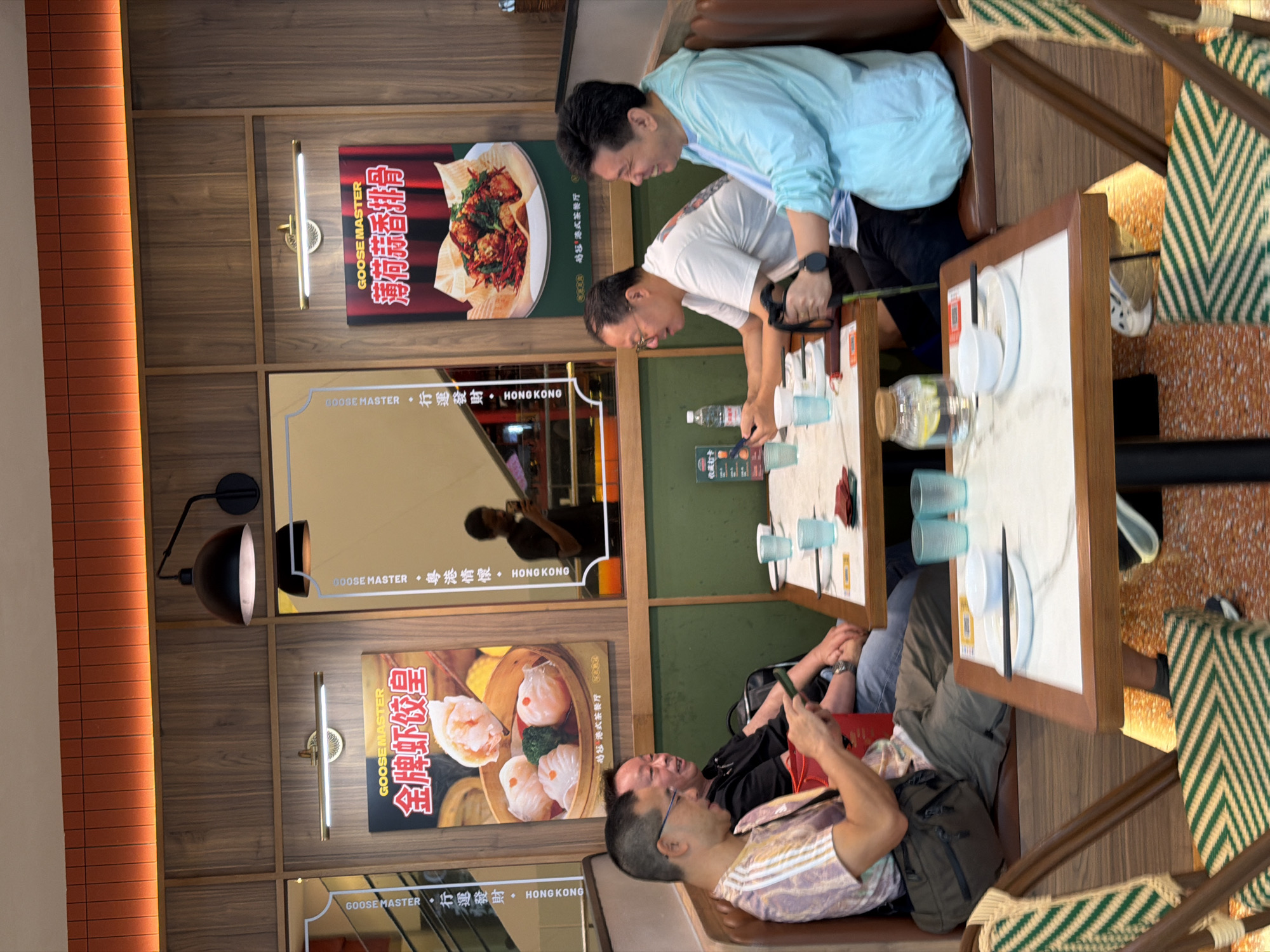 In the Yuzhong District, China, an indoor restaurant scene depicts four middle-aged Asian men seated at two tables. The ambiance is contemporary, with wooden paneling on the walls and orange lighting strips above. Two large posters advertising "GOOSE MASTER" food are prominently displayed. One poster features dumplings, and the other shows a dish of what appears to be fried ribs.
At the table closest to the viewer, two men are seated on the left. The man in the foreground, wearing a patterned shirt and khaki pants, is looking down at his phone. To his right, a man in a black shirt and jeans is looking to his right and smiling. On the table in front of them are blue glasses, plates, and chopsticks.
At the table further back, two men are seated on the right. The man on the left, wearing a white t-shirt, is looking to his left. The man on the right, wearing a light blue jacket, is also looking to his left and appears to be speaking. This table also has blue glasses and plates.
The overall environment suggests a casual dining experience with a focus on Chinese cuisine. The lighting is warm, contributing to a comfortable atmosphere. A young man in a black t-shirt is visible in the background reflection in a mirror, also holding a phone. Chairs with green and white striped upholstery are positioned at the tables.