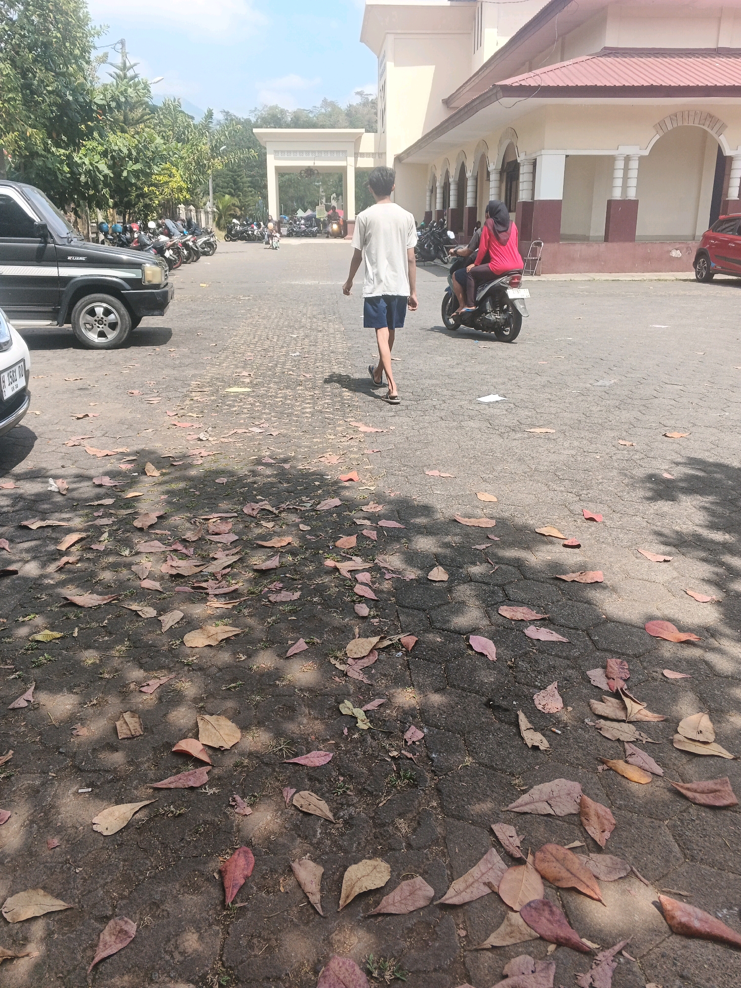 In the city of Kalirejo, Indonesia, an outdoor scene unfolds on a paved area littered with fallen leaves. In the foreground, a person walks away from the camera, wearing a white t-shirt and blue shorts. To their right, a motorcycle carrying two individuals, one wearing a red shirt and the other a hijab, travels away from the camera. Behind them, parked motorcycles and a black SUV are visible. A building with a red roof and light-colored walls, featuring arched windows and columns, forms the backdrop. The overall environment suggests a sunny day in a public space, possibly a parking lot or a courtyard, with a mix of natural elements and man-made structures.