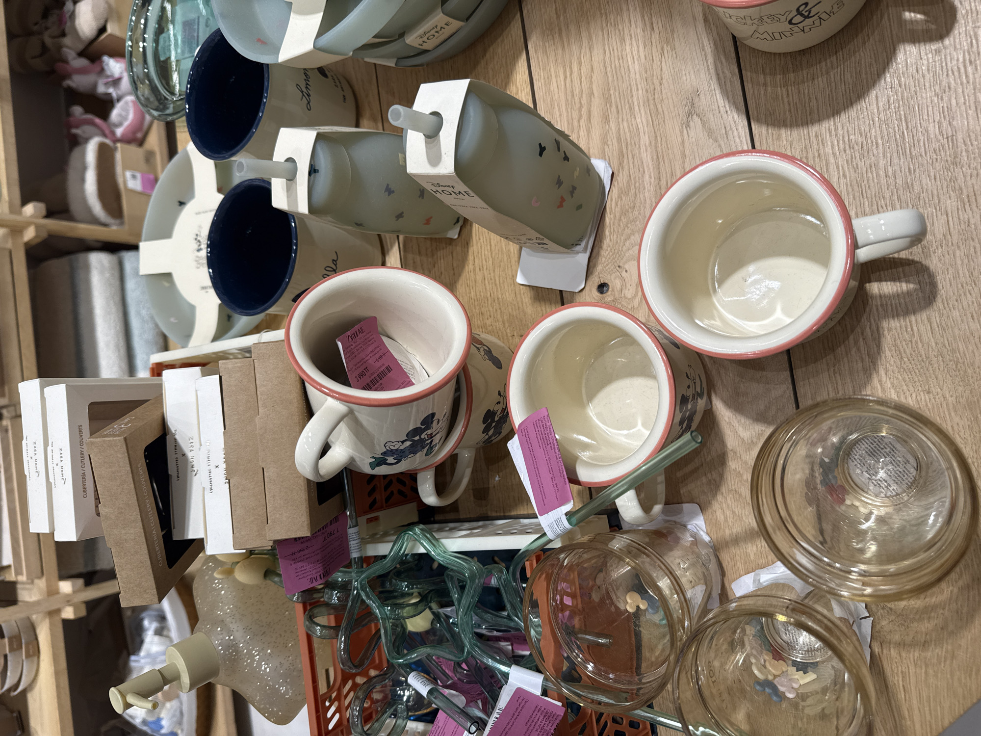 This overhead shot captures a retail display of homeware items, primarily mugs and glasses, arranged on a wooden surface. The items are predominantly white, cream, and pastel-colored. Several mugs are visible, some featuring Disney characters like Mickey and Minnie Mouse, with pink rims. There are also clear glass jars and tumblers, some containing small, colorful, flower-shaped items. A collection of green, star-shaped drinking straws is also present. Packaging boxes with "ZARA HOME" branding are scattered amongst the items. The scene appears to be inside a store, likely a home goods or department store, given the organized display and product packaging. There is no discernible activity or event taking place, as the image focuses on static products. The lighting suggests an indoor setting with artificial light. Visible text includes brand names like "ZARA HOME," "Disney," and "THE ANIMALS OBSERVATORY," along with price tags and item descriptions in what appears to be Cyrillic script, indicating the location is Almaty, Kazakhstan.