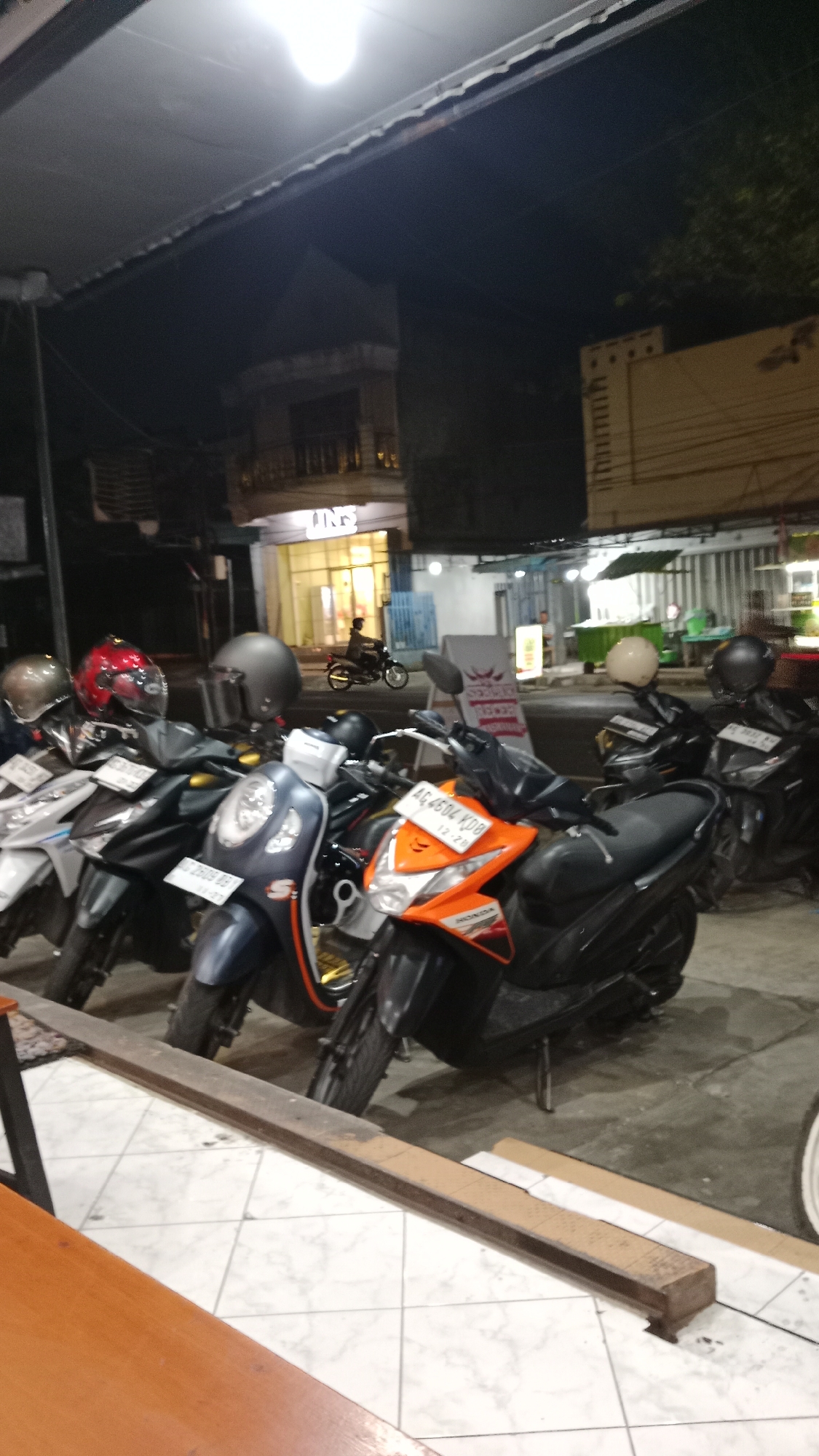 A nighttime exterior shot captures a row of parked motorcycles in front of a building in Blitar, Indonesia. The motorcycles are predominantly dark-colored scooters, with one orange Honda scooter prominent in the foreground. Several helmets are placed on the seats and handlebars of the motorcycles. In the background, a building with illuminated windows and signage is visible. A single person is seen riding a motorcycle in the distance, moving away from the camera. The ground is paved with checkered tiles in the foreground and asphalt in the background. Overhead, a bright light illuminates the scene.