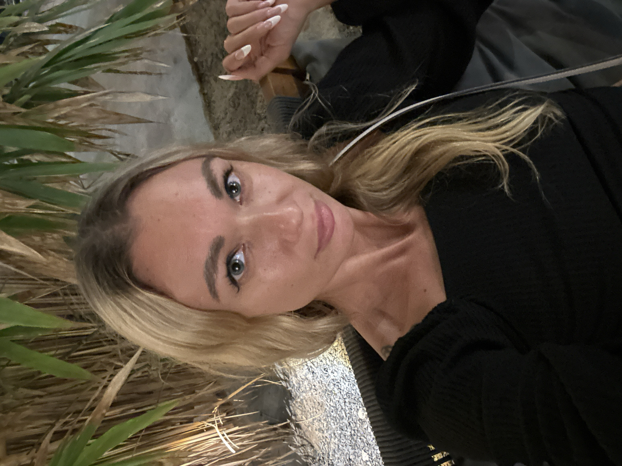 The image shows a selfie of a woman with blonde hair and blue eyes. She is wearing a black top with a square neckline and has long, light blonde hair that falls over her shoulders. Her nails are long and painted with a French manicure. She is sitting in what appears to be an outdoor seating area, possibly a patio or balcony, with a textured, dark-colored sofa or chair. In the background, there are tall, dry, grass-like plants and some green palm leaves, illuminated by artificial light. The ground appears to be covered in small pebbles. There is no visible indication of Phu Thuy Ward, Vietnam, in the image.