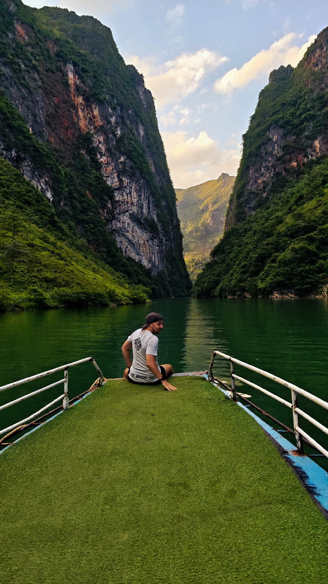 A man is sitting on the bow of a boat on a body of water in Đồng Văn Commune, Vietnam. The boat has a green astroturf deck and white railings. The man is facing away from the camera, looking back over his right shoulder. He is wearing a grey t-shirt, black shorts, and a baseball cap. The background consists of steep, green, and rocky canyon walls that extend upwards. The water in the river is green. There is a sky visible at the end of the canyon with light clouds.