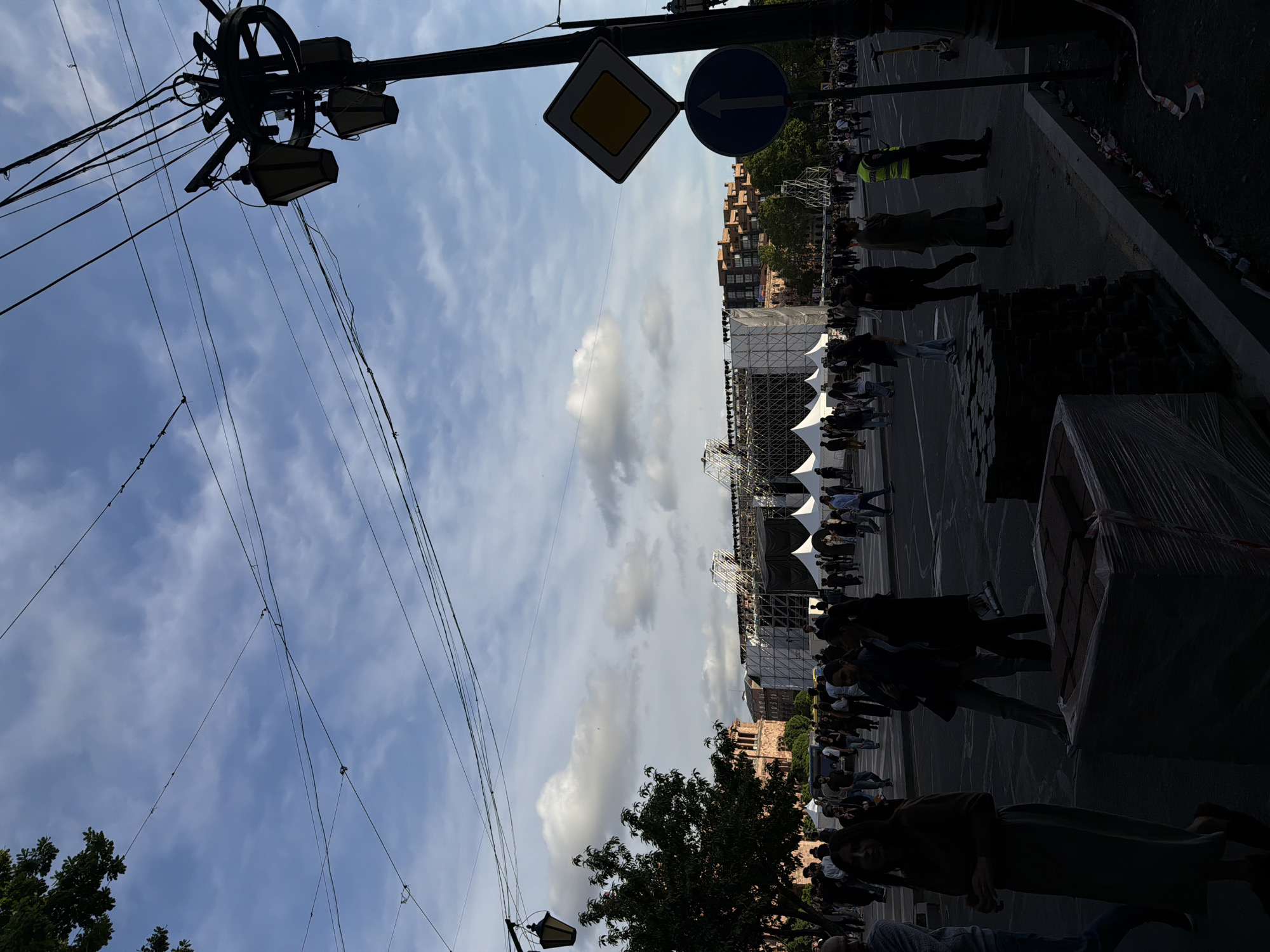 The image shows a street scene in Yerevan, Armenia. The view is angled upwards, capturing power lines, streetlights, and traffic signs. In the background, a stage is set up with a large crowd of people. People are walking around the stage. The weather is partly cloudy.