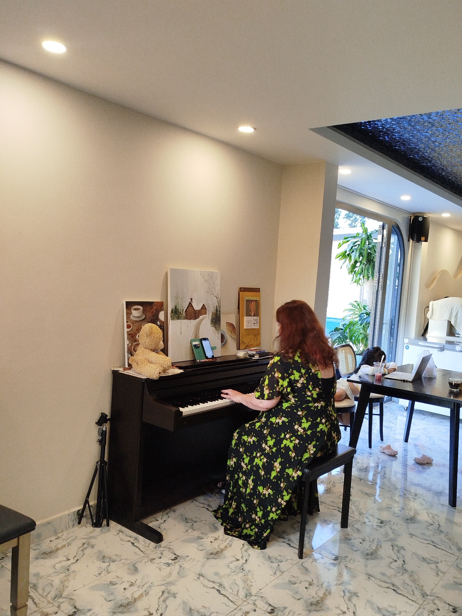 A woman with red hair sits at a black piano in a well-lit room, playing the instrument. She wears a dark, floral-patterned dress. On the piano, there are two framed pictures, a brown box, two cell phones and a stuffed bear. To the left of the piano stands a black tripod. The floor has a white marble pattern. Adjacent to the piano, a black table holds a laptop, a drink with a straw, and a glass. A young person sits at the table. The room opens to an outdoor area with plants and greenery. The location could possibly be Da Lat, Vietnam.