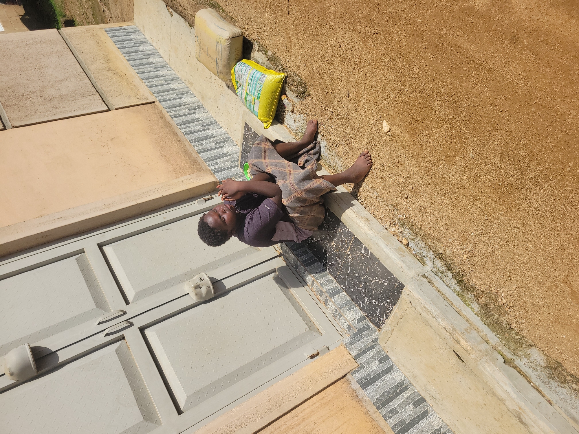 A young person with dark skin and short, curly hair is sitting on a concrete curb in front of a building with a light beige, textured facade. The building has a large, gray metal double door with diamond-patterned panels and two silver handles. To the right of the door, there are two large bags on the ground, one yellow and one pale yellow. The yellow bag has text and images printed on it. Near the yellow bag, there is a clear plastic bottle. The ground is covered in light brown, sandy soil. The person is wearing a dark purplish-brown t-shirt and plaid pants in shades of brown and gray. Their hands are clasped in front of their face, and they are looking downwards. The scene appears to be outdoors, under daylight. The presence of the building, the sandy ground, and the general aesthetic are consistent with a location in Jos, Nigeria.