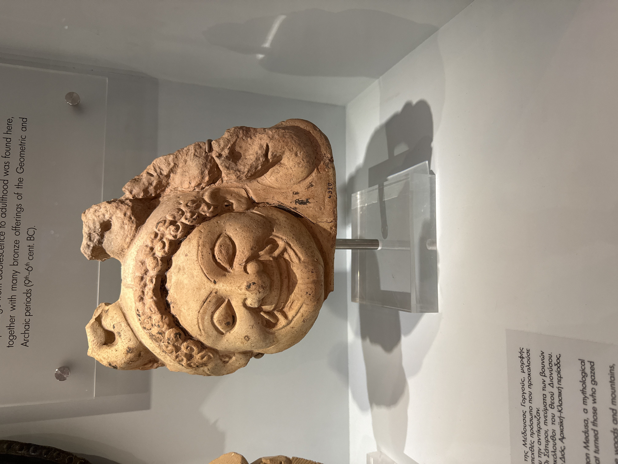 The image displays a terracotta architectural fragment, possibly a gorgoneion, at the Heraklion Archaeological Museum in Heraklion, Greece. The fragment depicts a grotesque, smiling face with wide eyes and elaborate hair or headdress. It is mounted on a clear acrylic stand against a plain, light-colored wall, creating a stark shadow on the surface behind it. Text labels are visible below and to the left of the artifact, indicating it is part of a museum exhibition. The lighting is even, illuminating the artifact and its display. The fragment has a weathered, earthy texture, characteristic of ancient terracotta. A number "4990" is faintly visible on the artifact's lower right side.
