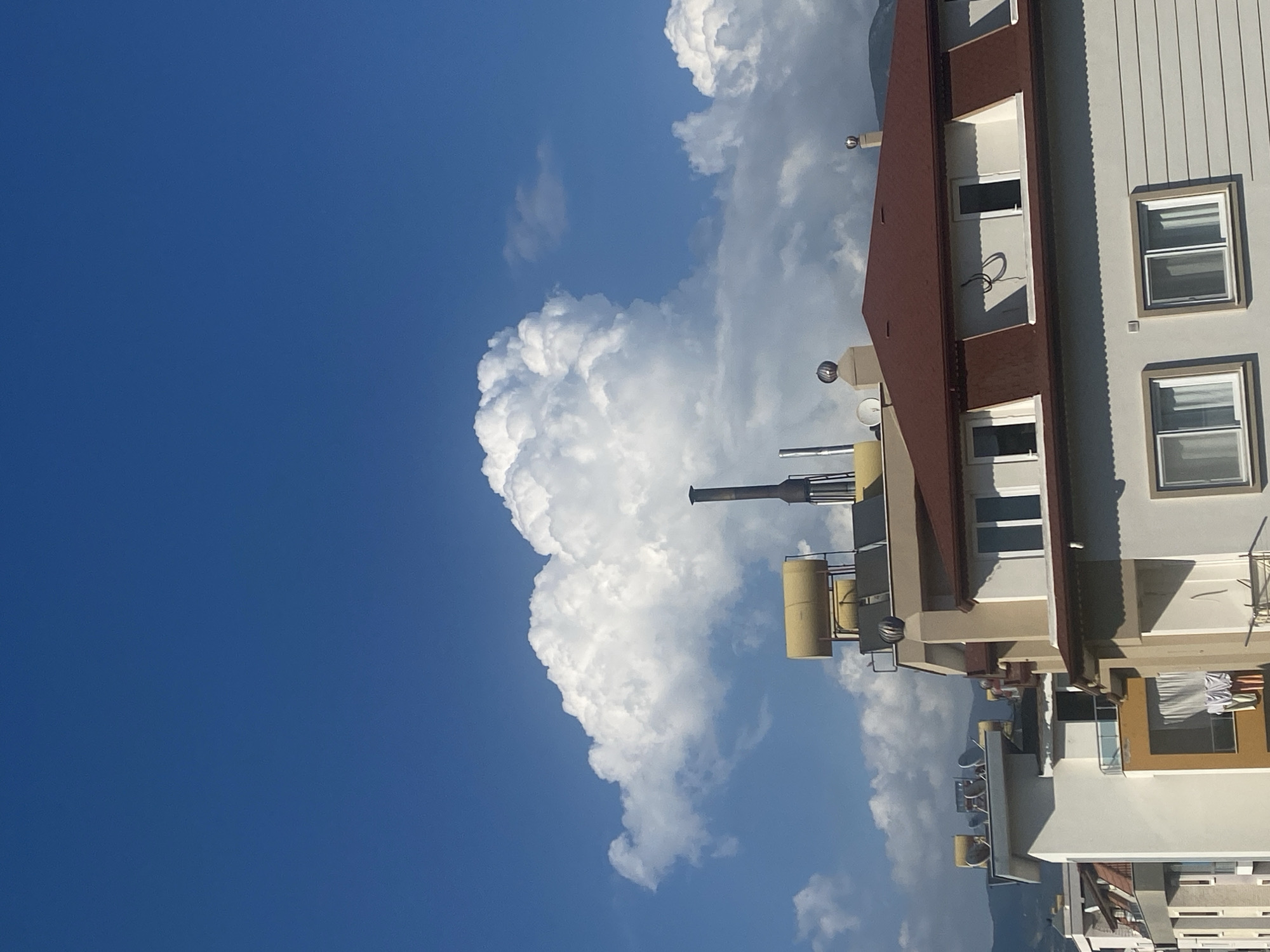 In Alanya, Turkey, a partially constructed building stands against a bright blue sky, dotted with large, fluffy white clouds. The building, seen from a slightly elevated angle, features a prominent, dark red tiled roof with dormer windows. The walls are painted in light colors, some with horizontal siding. On the rooftops of adjacent buildings, solar water heaters and satellite dishes are visible. A tall, dark chimney rises from one of the rooftops. The scene suggests a sunny day, possibly late morning or early afternoon, with a partly cloudy sky. The surrounding environment appears to be an urban or semi-urban area, with other buildings and distant hills or mountains visible in the background.