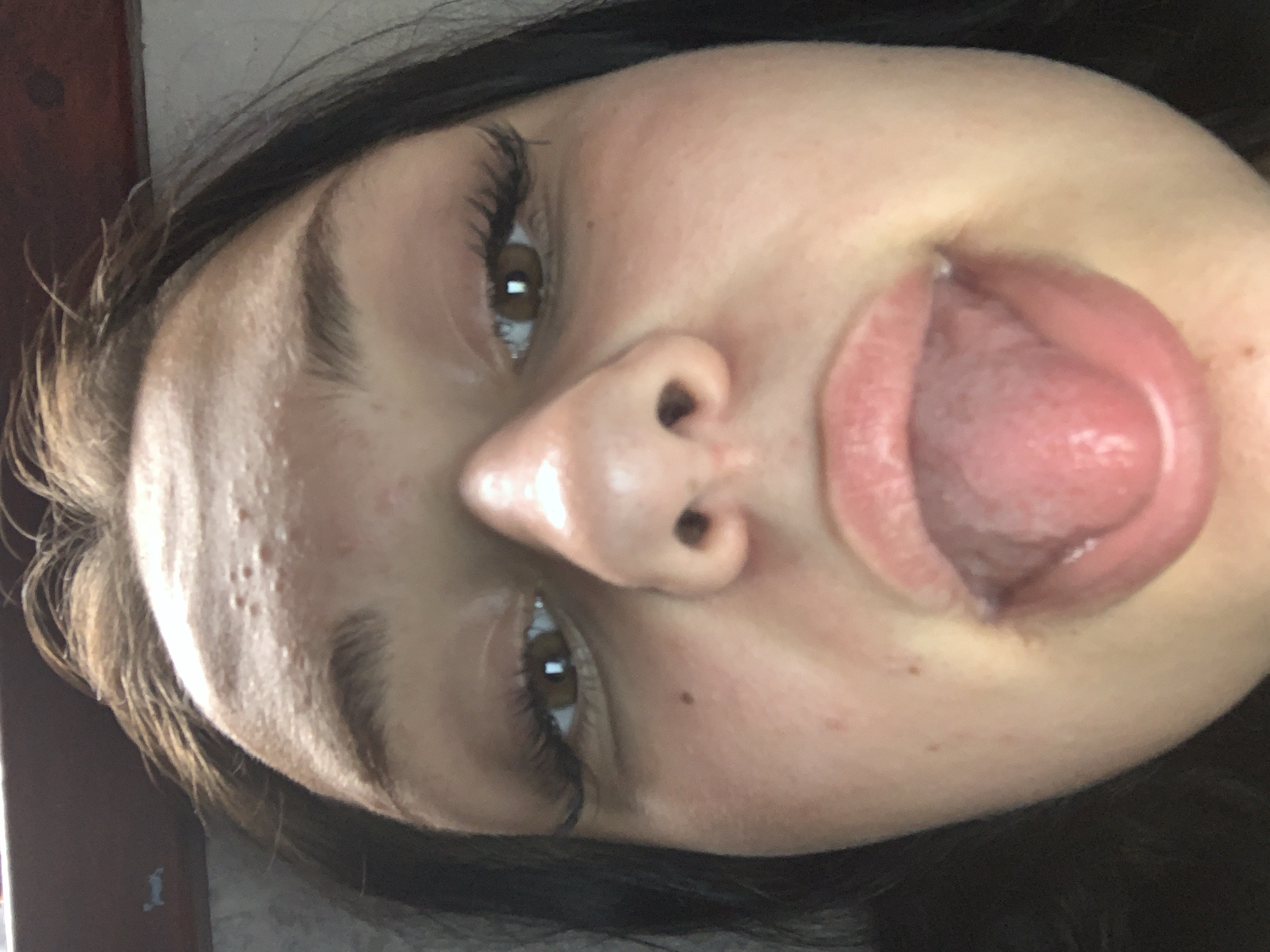 The image shows a close-up, low-angle selfie of a young person's face. The subject has light brown hair, brown eyes, and fair skin with some blemishes. Their tongue is extended out of their mouth. The background appears to be indoors, possibly near a window or shelf with a dark brown surface. The image does not contain any information about the city of Ustyvytsia, Ukraine.