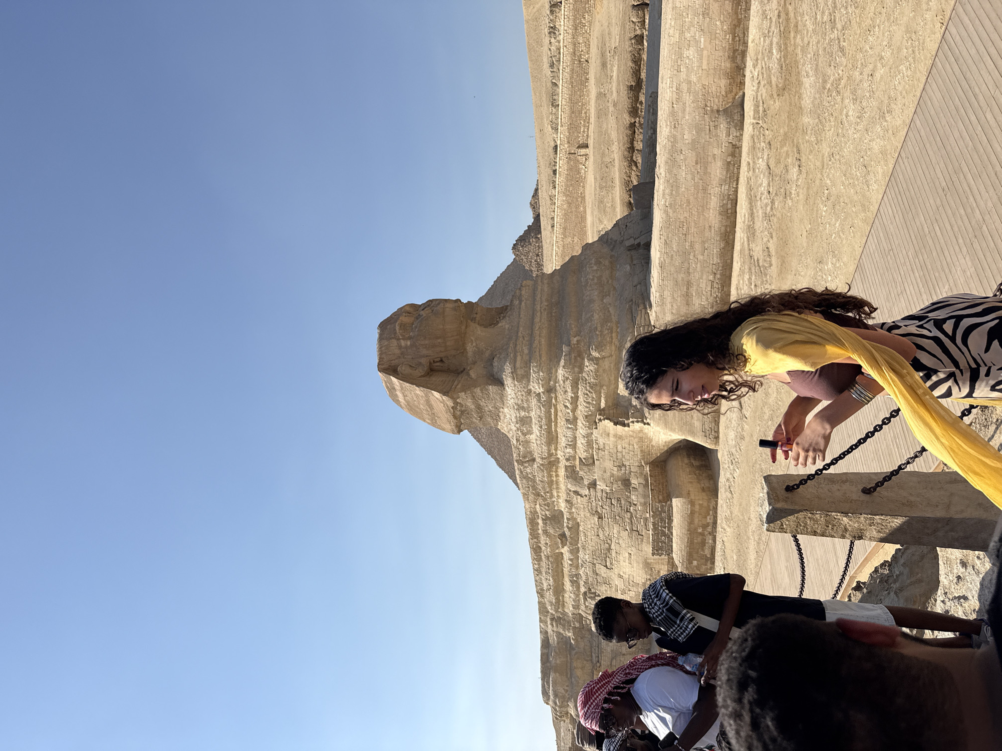 A group of tourists stand in front of the Great Sphinx of Giza, Egypt, on a clear, sunny day. The Sphinx, a limestone statue with the body of a lion and the head of a human, is partially obscured by a wooden walkway and a stone barrier in the foreground. Several people, including a woman wearing a yellow scarf and a zebra-print skirt, are looking towards the Sphinx. Another woman is holding a lipstick in her hand. Two men are wearing keffiyeh scarves. The surrounding landscape is sandy and arid, with stone structures and the Great Pyramid of Giza visible in the background.